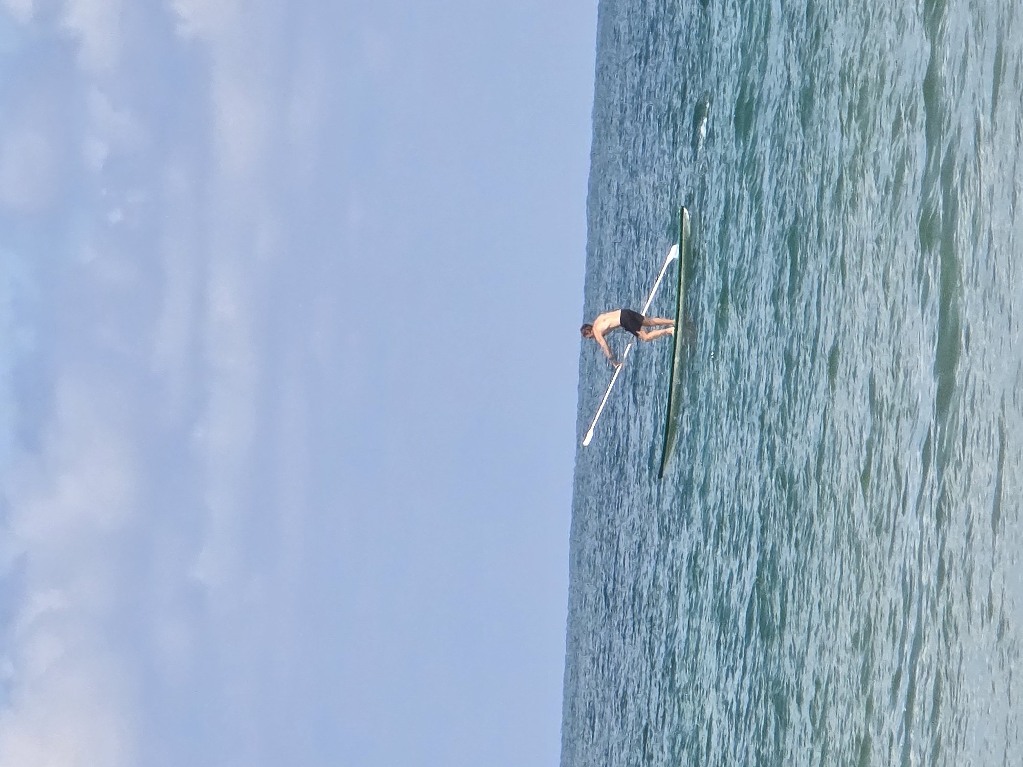 A man rows a single scull on the Mediterranean Sea off the coast of Tel Aviv, Israel. The sky is blue with some scattered clouds. The water is turquoise and choppy. The man is wearing dark shorts and rowing with oars.