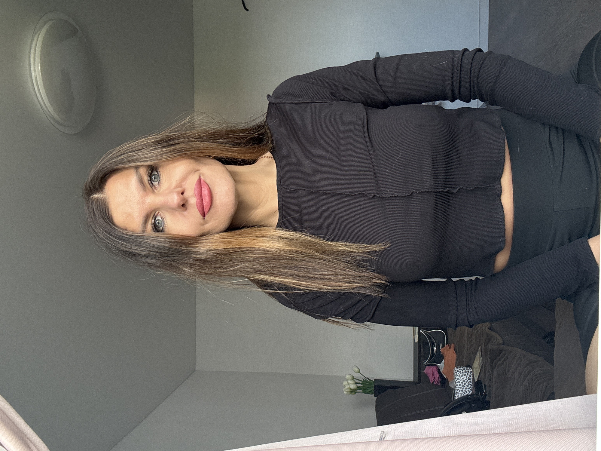 A woman with long brown hair and fair skin poses for a photo indoors. She is wearing a black long-sleeved crop top and black pants. Her gaze is directed towards the camera, and she has light blue eyes and pink lips. The background is a room with light gray walls. A circular light fixture is visible on the upper left wall. A bouquet of white tulips is positioned near the bottom center of the frame. The lower portion of the photo reflects a sofa, chair, and other objects in a mirror. The location is presumed to be an interior space in a city of Chaikovsky, Russia.