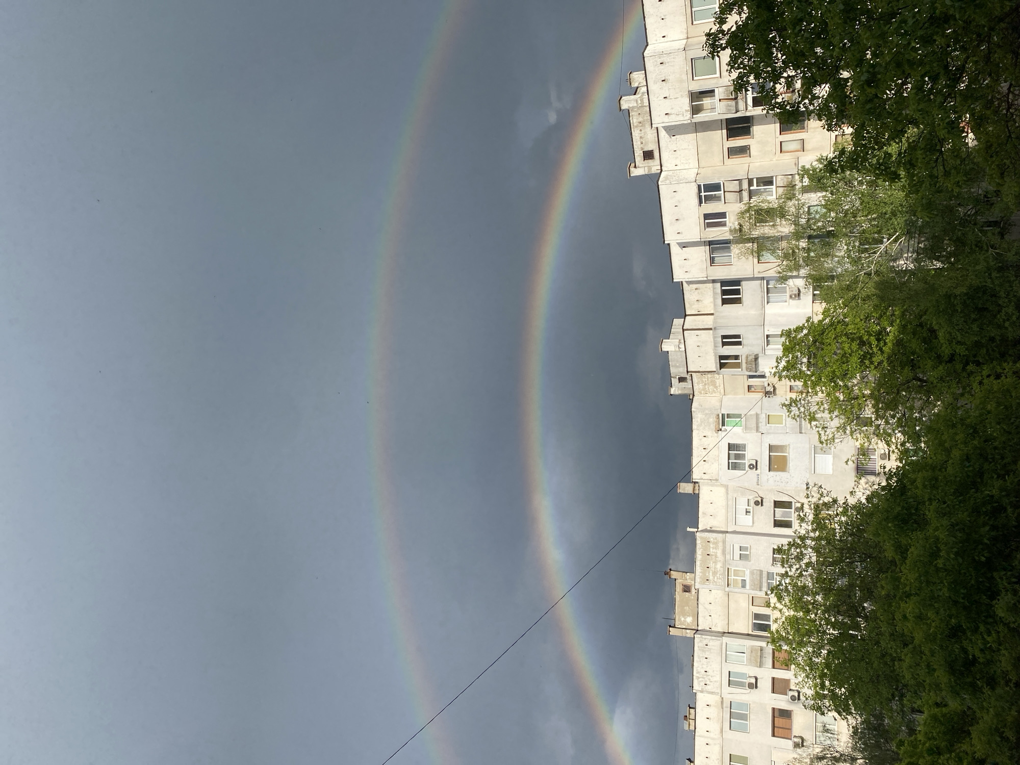 The image captures a double rainbow in the sky above Pleven, Bulgaria. The sky is overcast with dark gray clouds. Adjacent to the rainbow is a tall, concrete apartment building with numerous windows and air conditioning units. Green trees partially obscure the building on the right side of the frame. A thin power line bisects the scene diagonally.