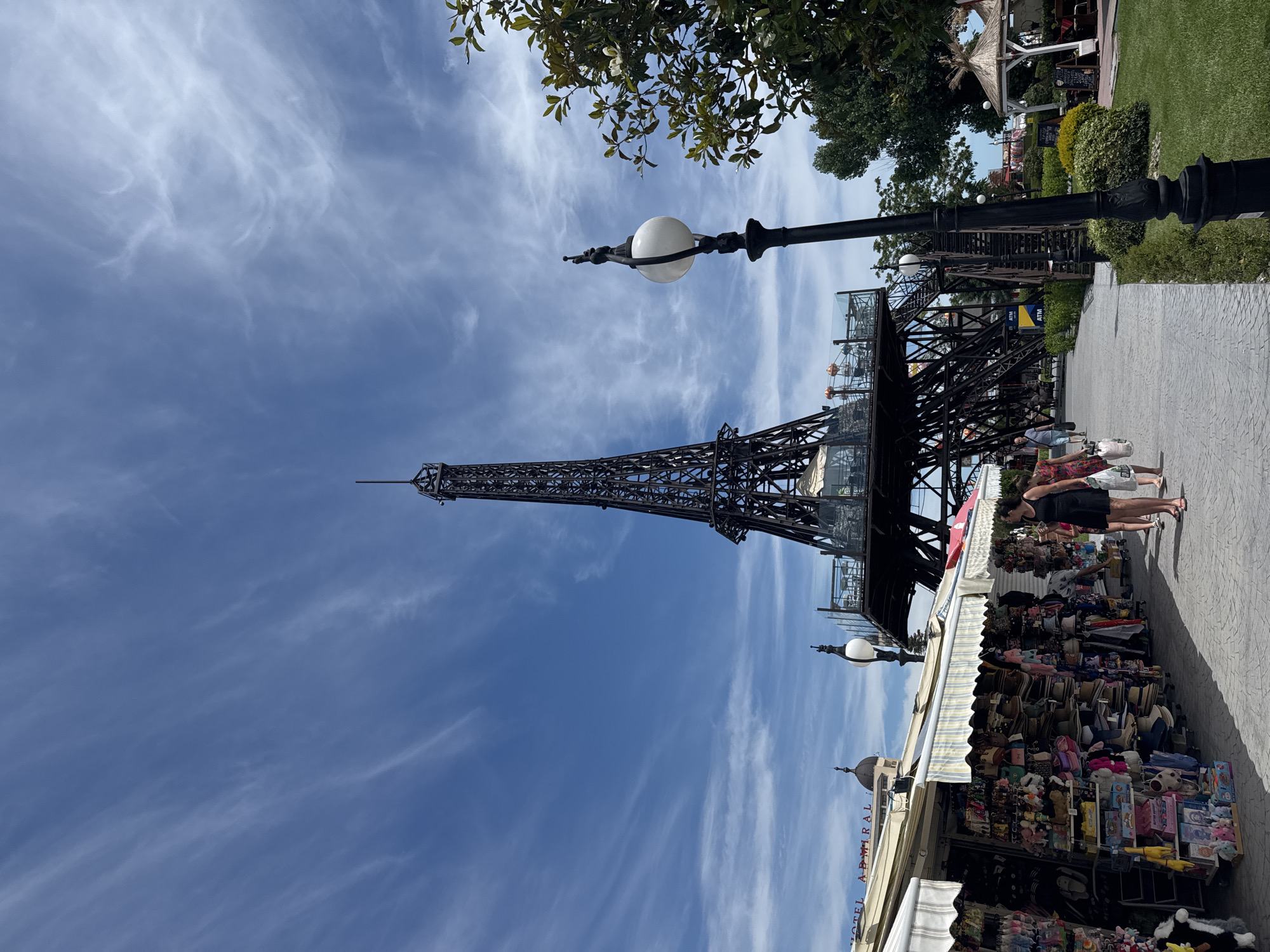 The image shows a replica of the Eiffel Tower located in the city of Varna, Bulgaria. The tower is made of black metal and has a viewing platform with tables and chairs. In front of the tower, there is a promenade with shops selling souvenirs and a few people walking. The sky is blue with scattered clouds. The replica Eiffel Tower is situated in a park-like area with green grass and trees. The overall scene is a tourist attraction with commercial activities.