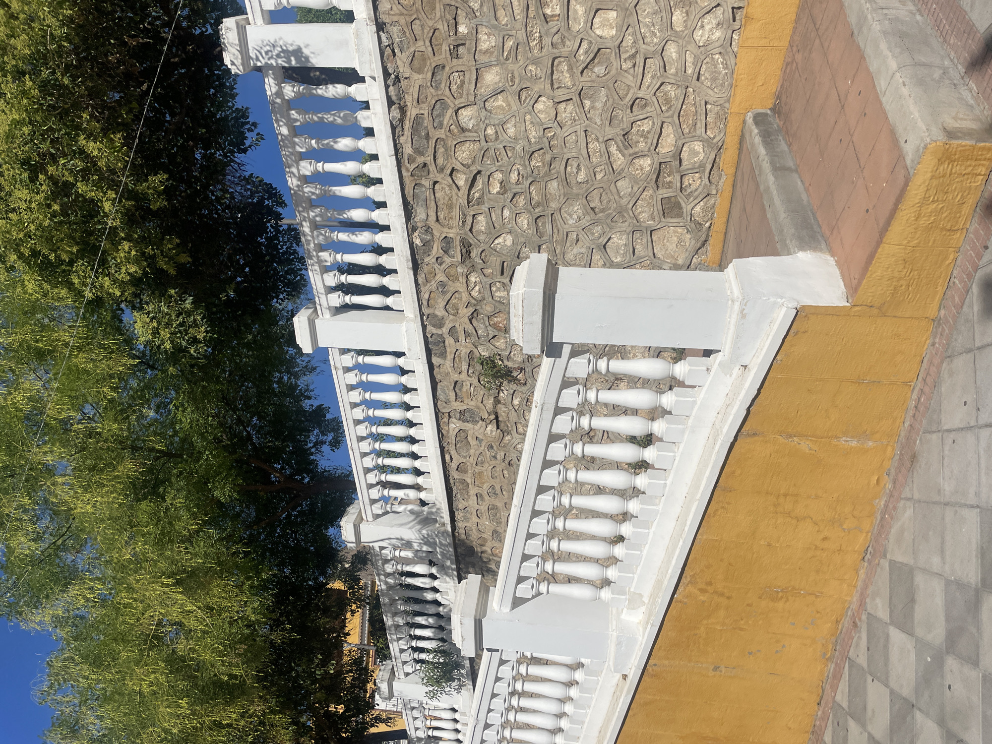 The image displays a view of a white, ornate balustrade with decorative posts, ascending a set of stairs. The stairs are bordered by a vibrant yellow wall. Behind the balustrade, a stone wall is visible, constructed from irregularly shaped stones. Above the stone wall, a lush green tree with drooping branches and small, elongated seed pods can be seen against a clear blue sky. To the left, a portion of a yellow building is partially obscured by the tree. The foreground shows a checkered pavement pattern. The scene is situated in the city of Atarfe, Spain, specifically on the Rambla de Béznar street. The lighting suggests it is daytime, with shadows cast by the balustrade and trees. There are no people or animals present in the image.