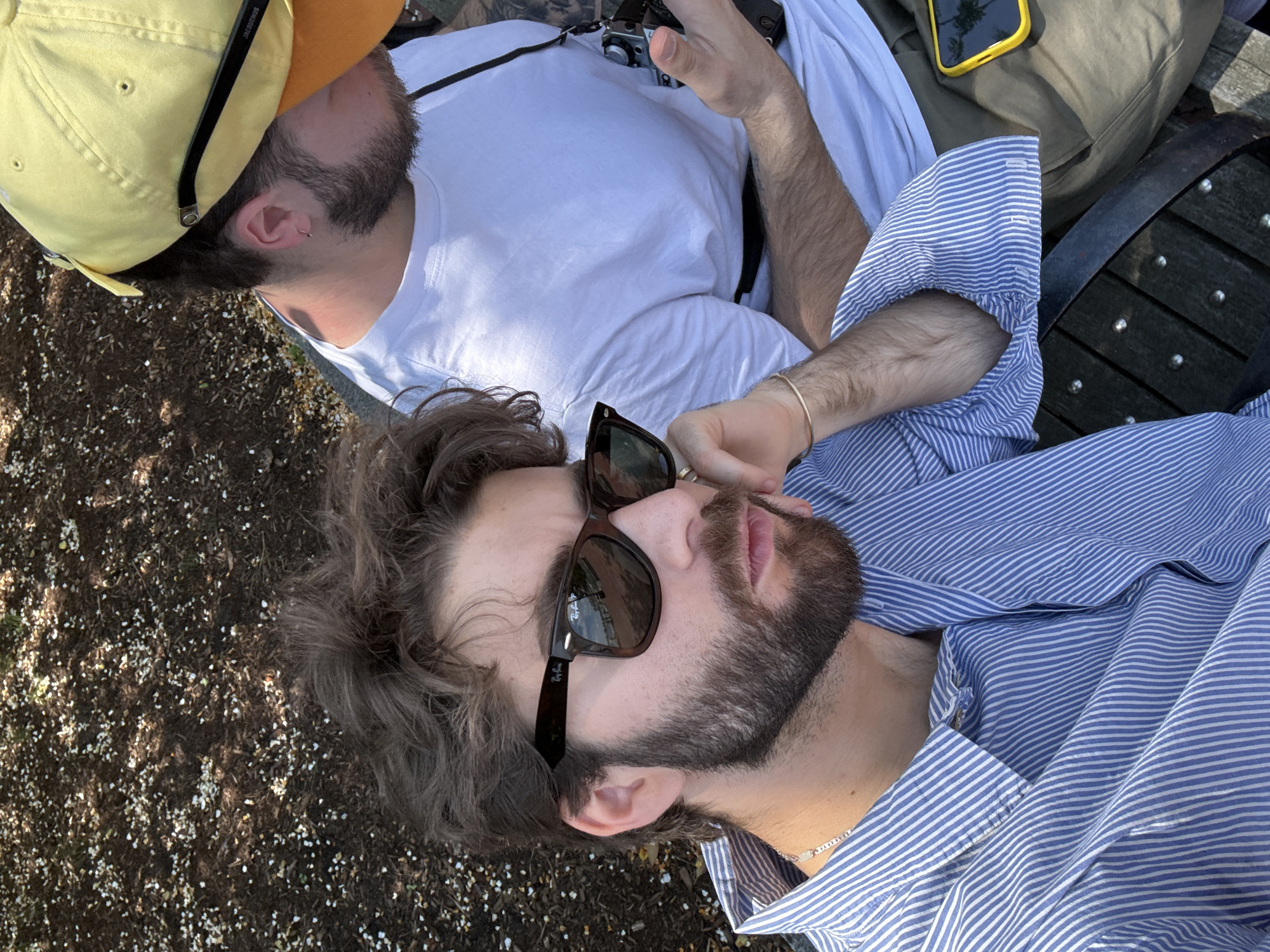 Two young men are reclining on an outdoor park bench, likely in Boston, USA. One man, wearing a yellow cap and a white t-shirt, is holding a camera and interacting with it. The second man is wearing a blue striped shirt and sunglasses. The background features ground covered with mulch and small white petals. A yellow cellphone rests between the two individuals.