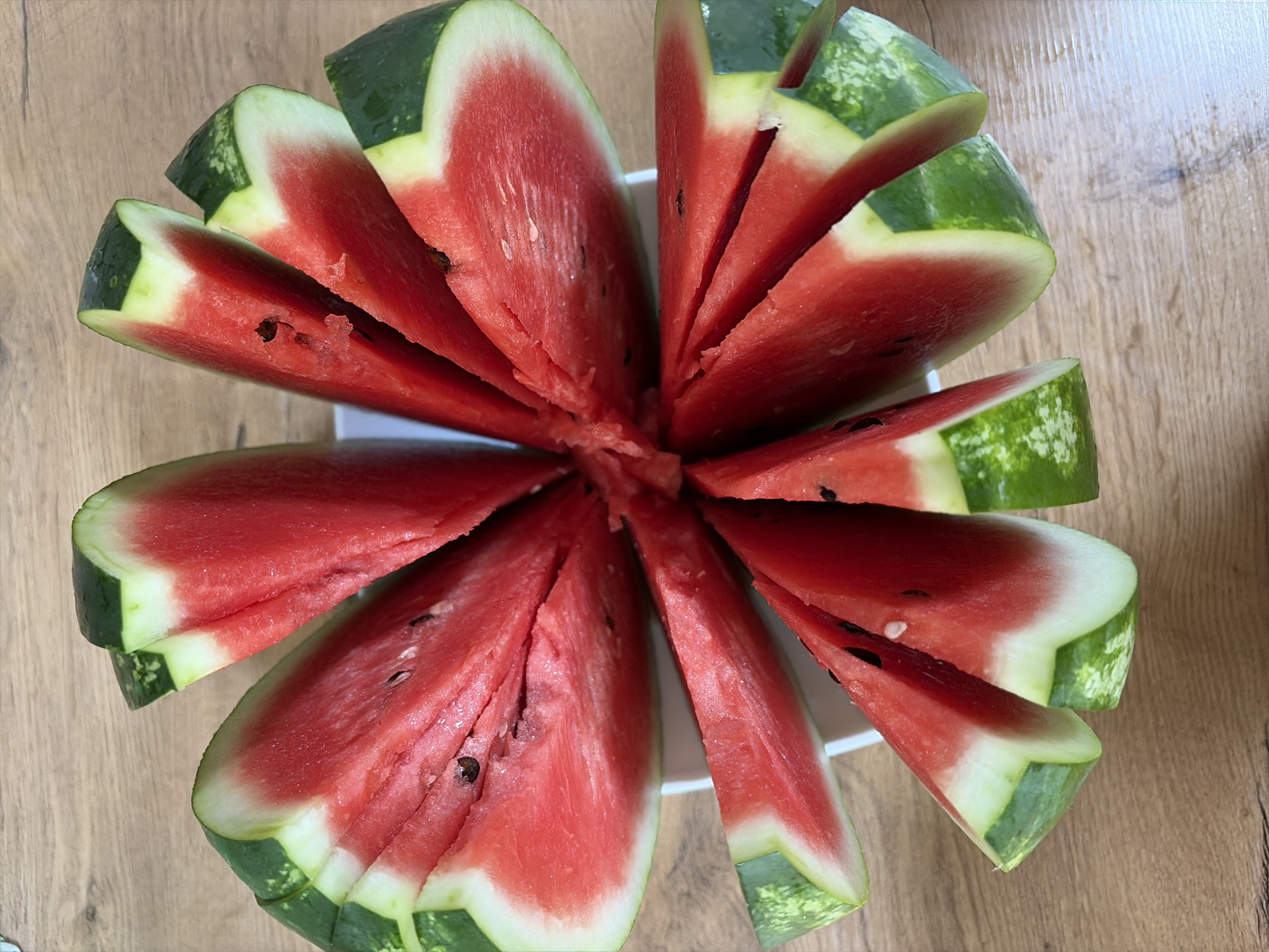 A whole watermelon has been cut into wedges and arranged in a circular pattern on a white plate, which is placed on a wooden surface. The watermelon slices are vibrant red with a white rind and a green outer peel. Some slices show black seeds. The lighting suggests an indoor setting, possibly a kitchen or dining area. There are no people or animals present in the image. The scene is static and appears to be a simple arrangement of cut fruit.