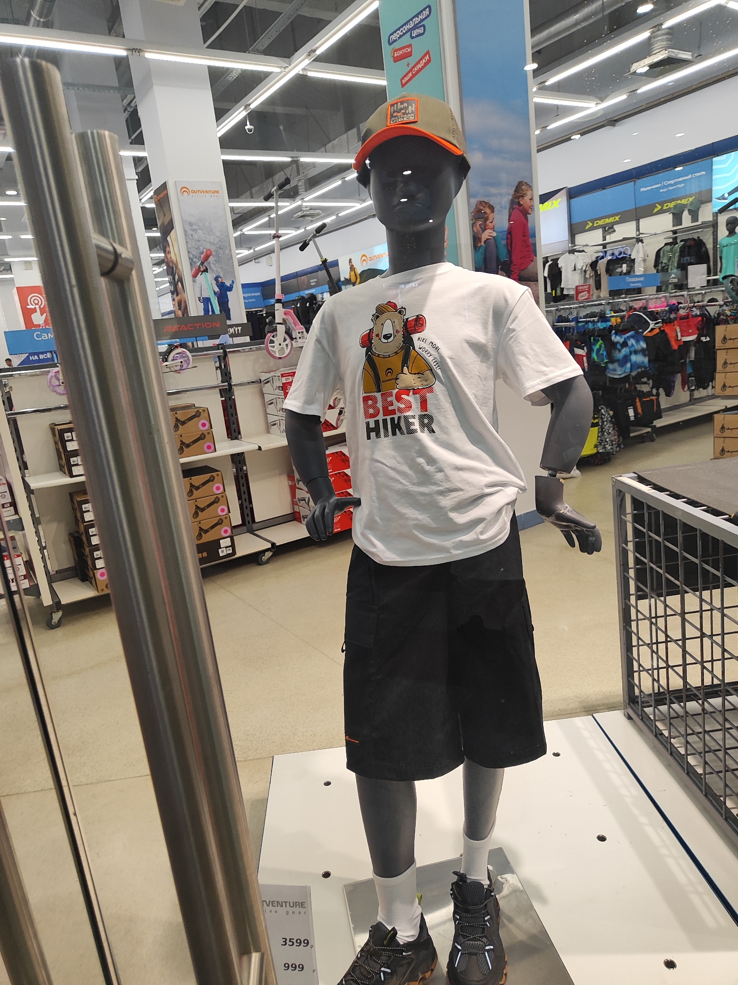 In a retail store, a male mannequin is displayed wearing a white t-shirt with a graphic of a bear and the text "BEST HIKER", black shorts, white socks, and black athletic shoes. The mannequin also wears an orange and grey baseball cap. The background shows shelves stocked with clothing and merchandise, as well as promotional posters. To the left of the mannequin, a tall, metallic display pole is visible.