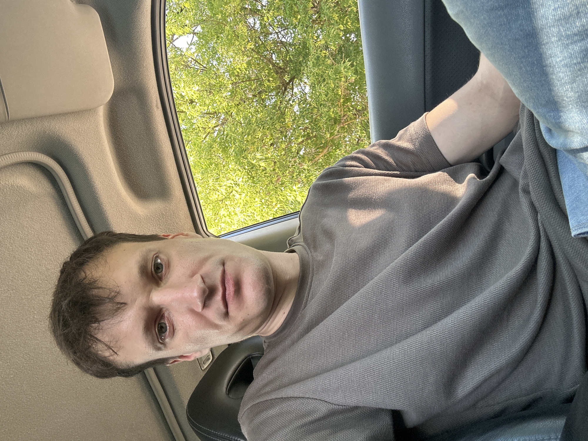 A man is taking a selfie inside a car. The man has brown hair and is wearing a grey t-shirt and blue jeans. He is looking directly at the camera with a neutral expression. Outside the car window, there are green trees. The interior of the car appears to be grey and beige. The image is taken during the daytime, as indicated by the natural light coming through the window. There is no visible indication of the location in the city of Makiv, Ukraine.