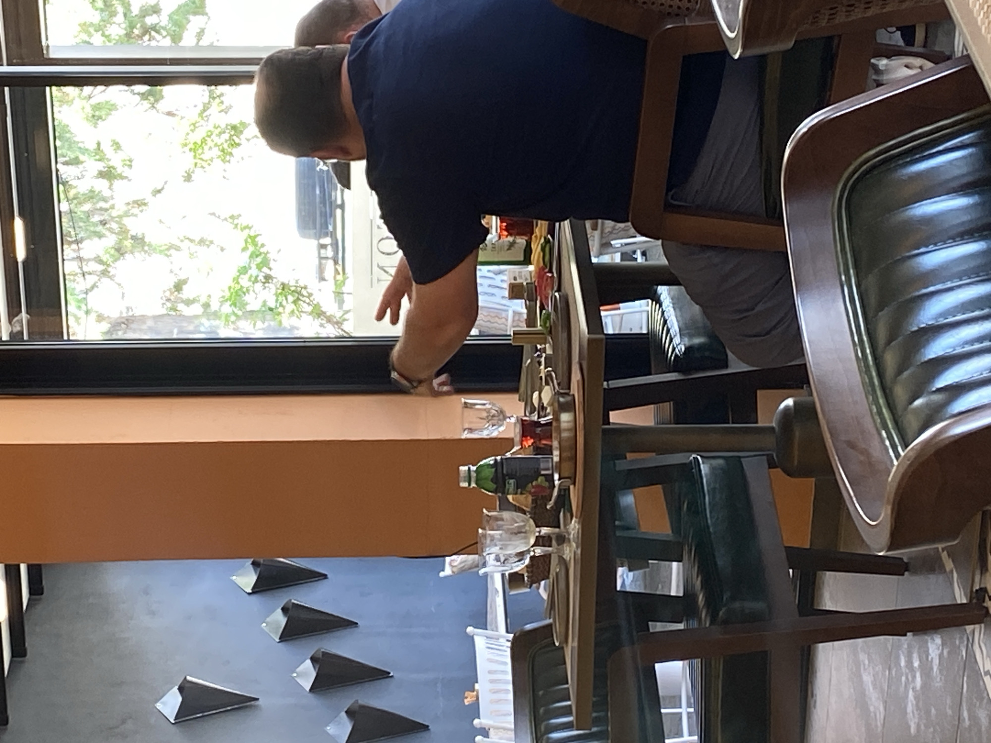 The image shows the interior of a restaurant in Sokhumi, Abkhazia. A man in a dark blue t-shirt is seated at a table, facing away from the camera. He is gesturing with his right hand. The table is set with plates, glasses, and a bottle of drink. In the background, there is a large window with trees visible outside. The walls are painted a dark blue and a light orange color, and there are decorative elements on the blue wall that resemble ice cream cones. The flooring appears to be tiled. Several chairs with dark green upholstery are around the table.