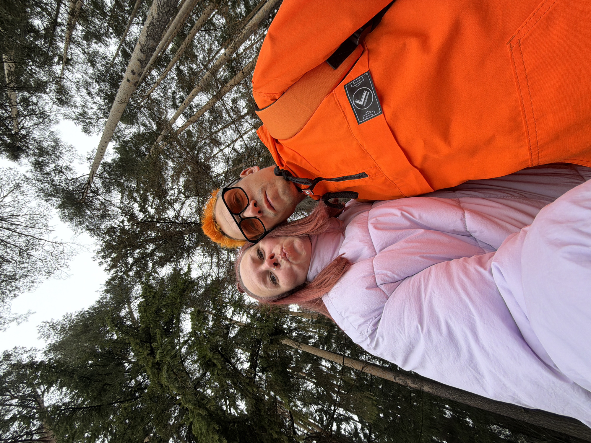 Two people lie on the ground in a forest, likely in the city of Chaikovsky, Russia, based on the image metadata (not provided). A man with dyed orange hair and sunglasses wears an orange jacket. A woman with dyed pink hair wears a light purple puffer jacket. They are positioned close together, and the photo is taken from a low angle, looking up toward the trees. Tree trunks and foliage fill the background. The sky is overcast.