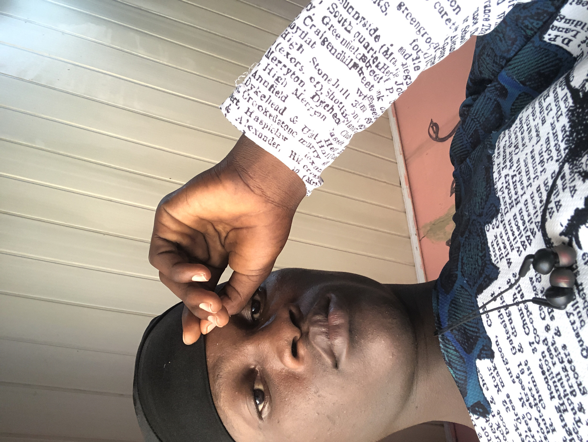 The image is a close-up, slightly angled selfie of a young Black man wearing a black durag and a blue and green patterned shirt with text print. He is looking towards the camera with his right hand touching his forehead. The background appears to be an indoor ceiling with white horizontal panels, possibly in a room in Ado, Nigeria. The man has dark skin, brown eyes, and is wearing black earbuds around his neck. The text on his shirt is small and appears to be a list of names and professions. The lighting suggests an indoor setting with natural light coming from the side.