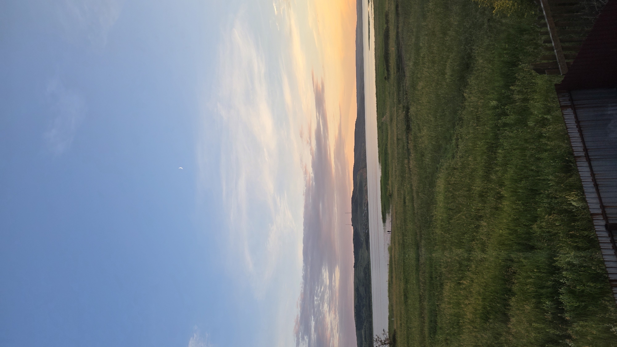 An outdoor, vertical, medium shot captures a sunset over a tranquil body of water. The sky transitions from a pale blue at the top to hues of pink, orange, and yellow near the horizon. A thin crescent moon is visible in the upper portion of the sky. In the middle ground, a wide body of water stretches across the frame, bordered by a distant, rolling, tree-covered hill. A small village or cluster of buildings is faintly visible on the hill. The foreground features a vast, grassy field with tall, green vegetation. At the very bottom of the frame, a weathered, dark metal fence runs horizontally, partially obscuring the lower edge of the field. Two small figures, appearing to be humans, are standing near the water's edge in the middle ground. The scene evokes a sense of rural serenity. The location is identified as Kupoyarovo, Russia.