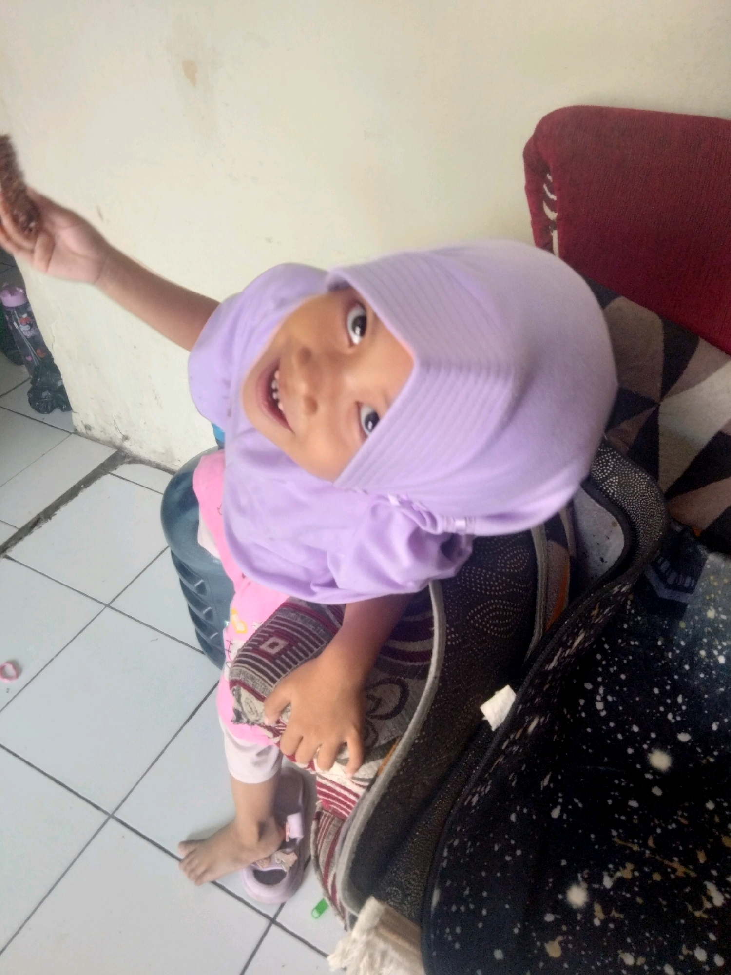 A young child wearing a purple hijab and pink pants sits on a patterned cloth, holding a brown object, possibly food, in their right hand. The child is looking up at the camera with a wide smile, revealing their teeth. Their bare left foot is visible, resting on a pink sandal. The background consists of a light-colored wall and a tiled floor. In the upper left corner, a pink water bottle with a patterned label can be seen. The scene appears to be indoors, likely in a residential setting in Suka Sari, Indonesia.