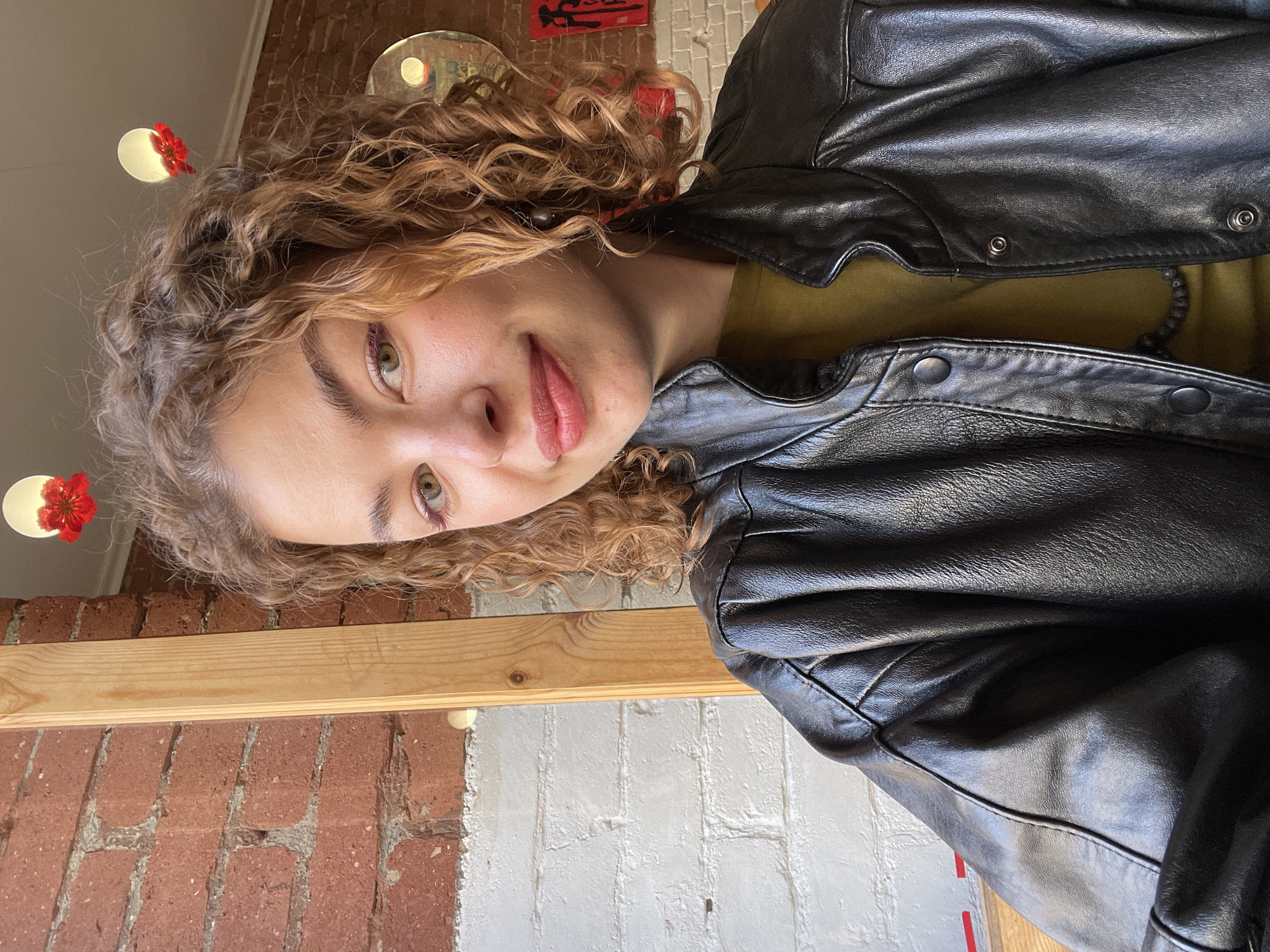 A close-up, tilted portrait features a young woman looking directly at the camera with a gentle, natural smile. She has voluminous, curly light brown hair and light skin, with hazel-green eyes and subtle makeup. She is dressed in a black faux leather jacket, worn over an olive-green top, with a dark, possibly beaded necklace visible.
The scene is indoors, characterized by architectural textures. The background shows a wall with sections of exposed red brick and segments of painted white brick, suggesting a renovated space or cafe setting. A horizontal wooden beam stretches across the middle ground. Above, circular light fixtures are visible, each adorned with a small red floral embellishment. A reflective, circular metallic object, possibly a decorative mirror or plate, is partially seen in the upper background, reflecting one of the lights.
The lighting is soft and diffused, illuminating her face and the immediate surroundings. The overall emotion conveyed is calm and pleasant. On a red rectangular sign in the upper background, dark, stylized characters, possibly East Asian, are faintly discernible but not explicitly readable. There are no other visible texts or clear indicators of specific activities beyond posing for the photograph. The image, consistent with interior settings found in Tbilisi, Georgia, captures a moment of serene contemplation or a casual portrait.