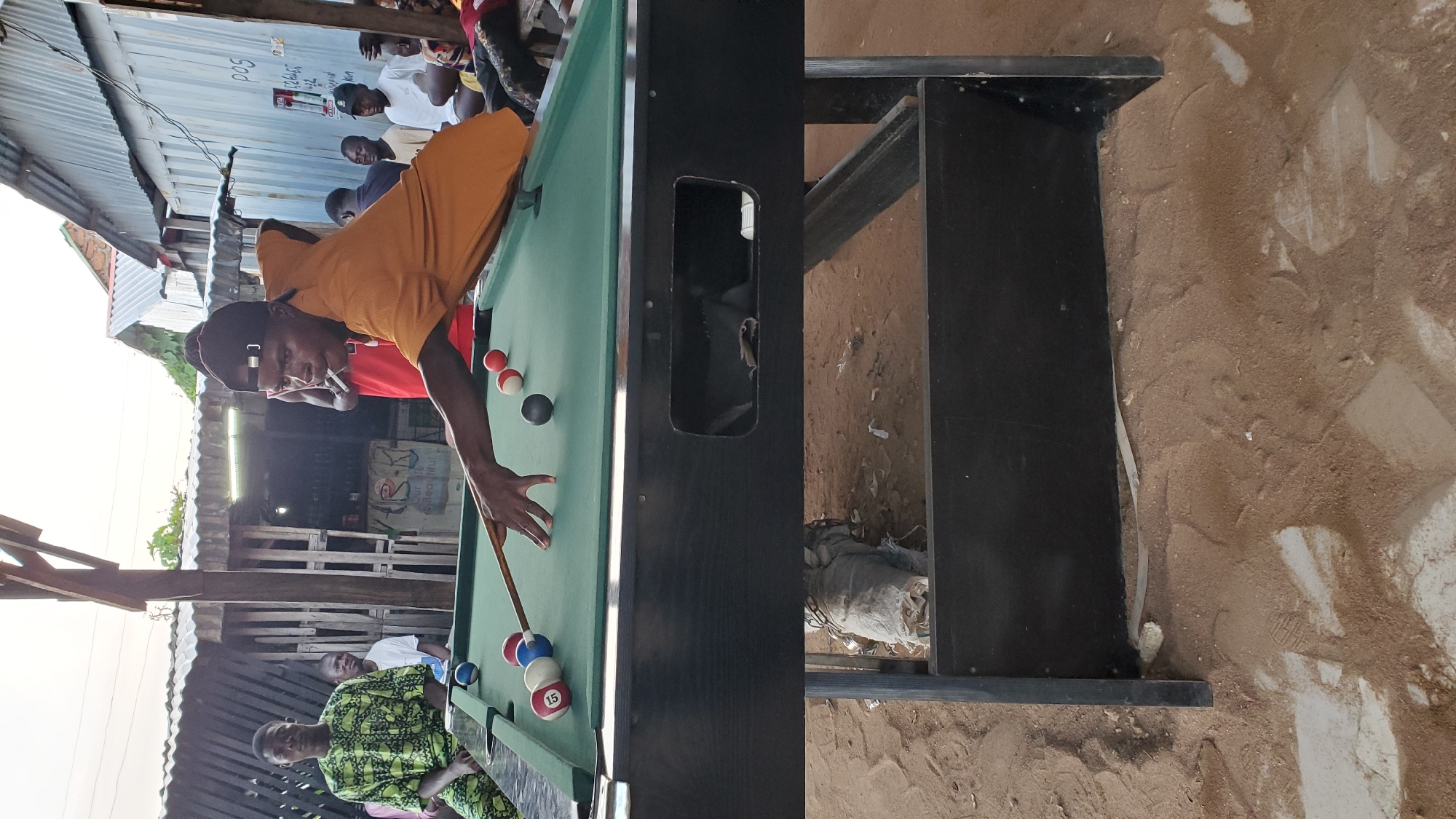 An outdoor pool table scene in Jalingo, Nigeria, shows several individuals engaged in or observing a game of pool. The table is set on uneven, sandy ground with scattered debris. A young man wearing a dark baseball cap and an orange t-shirt is actively playing, holding a cue stick and positioned over the table. He is smoking a cigarette. Several pool balls, including a striped ball numbered "15", a blue ball, and a red ball, are on the green felt surface. In the background, other men are visible, some seated and others standing, watching the game or socializing. They are dressed in casual attire, including a man in a patterned green and black dashiki. The setting appears to be a casual, open-air establishment, with corrugated metal structures and rudimentary fencing visible behind the players, suggesting a local community gathering spot. The lighting suggests it is late afternoon or early evening.