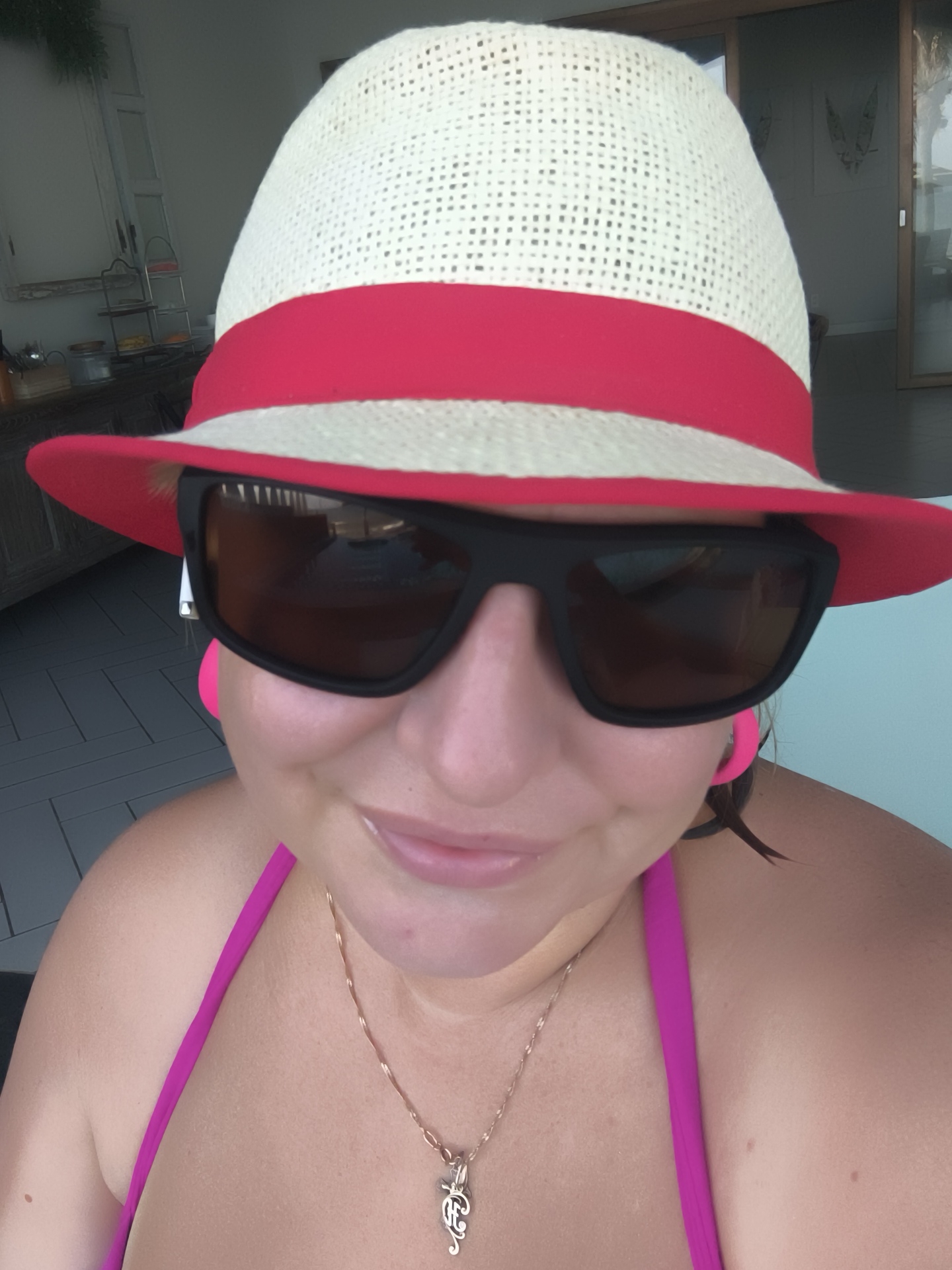 A woman is posing for a selfie wearing a white and red striped straw hat, large black sunglasses, and a pink bikini top. She is also wearing a gold necklace with a pendant. The background appears to be an outdoor setting, possibly a patio or balcony, with a view of a modern building with large glass doors and some greenery. The lighting suggests it is daytime. The specific location in Phu Thuy Ward, Vietnam cannot be determined from the image alone.