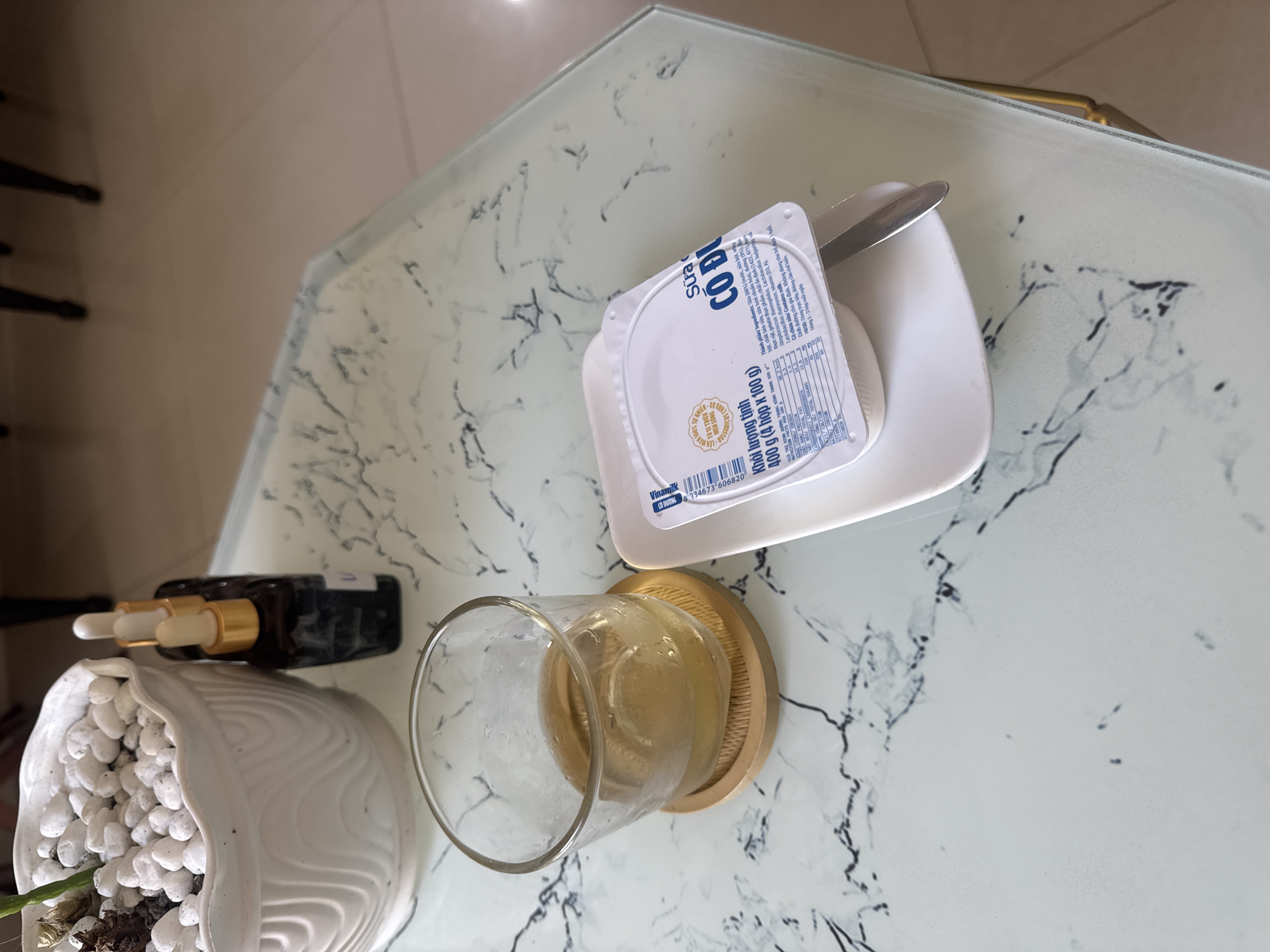 The image displays a top-down view of items arranged on an octagonal glass table with a white marble-patterned surface. The location is identified as Nha Trang, Vietnam, based on the visible "Vinamilk Sữa Chua Có Đường" (Sweetened Yogurt) product, a prevalent Vietnamese brand.
In the upper left, a white ceramic planter with a ridged texture contains white decorative pebbles and a plant stem. Immediately behind this planter, three small, dark amber glass dropper bottles with gold-colored caps and white droppers are positioned on the table.
Near the center-left, a clear glass holding a light amber liquid, consistent with tea, rests on a round, light brown bamboo coaster. Condensation is present on the exterior surface of the glass.
On the right side of the frame, a white rectangular ceramic saucer supports a white plastic container of Vinamilk Sweetened Yogurt. A silver-colored spoon is placed on the right edge of the yogurt container. The label indicates "Khối lượng tịnh: 400 g (4 hộp x 100 g)" (Net weight: 400 g (4 cups x 100 g)).
The table's right edge features a metallic trim. The blurred background shows a light brown tiled floor and the dark, indistinct legs of chairs, suggesting an indoor setting such as a cafe, home, or hotel room. No people or active interactions are depicted.