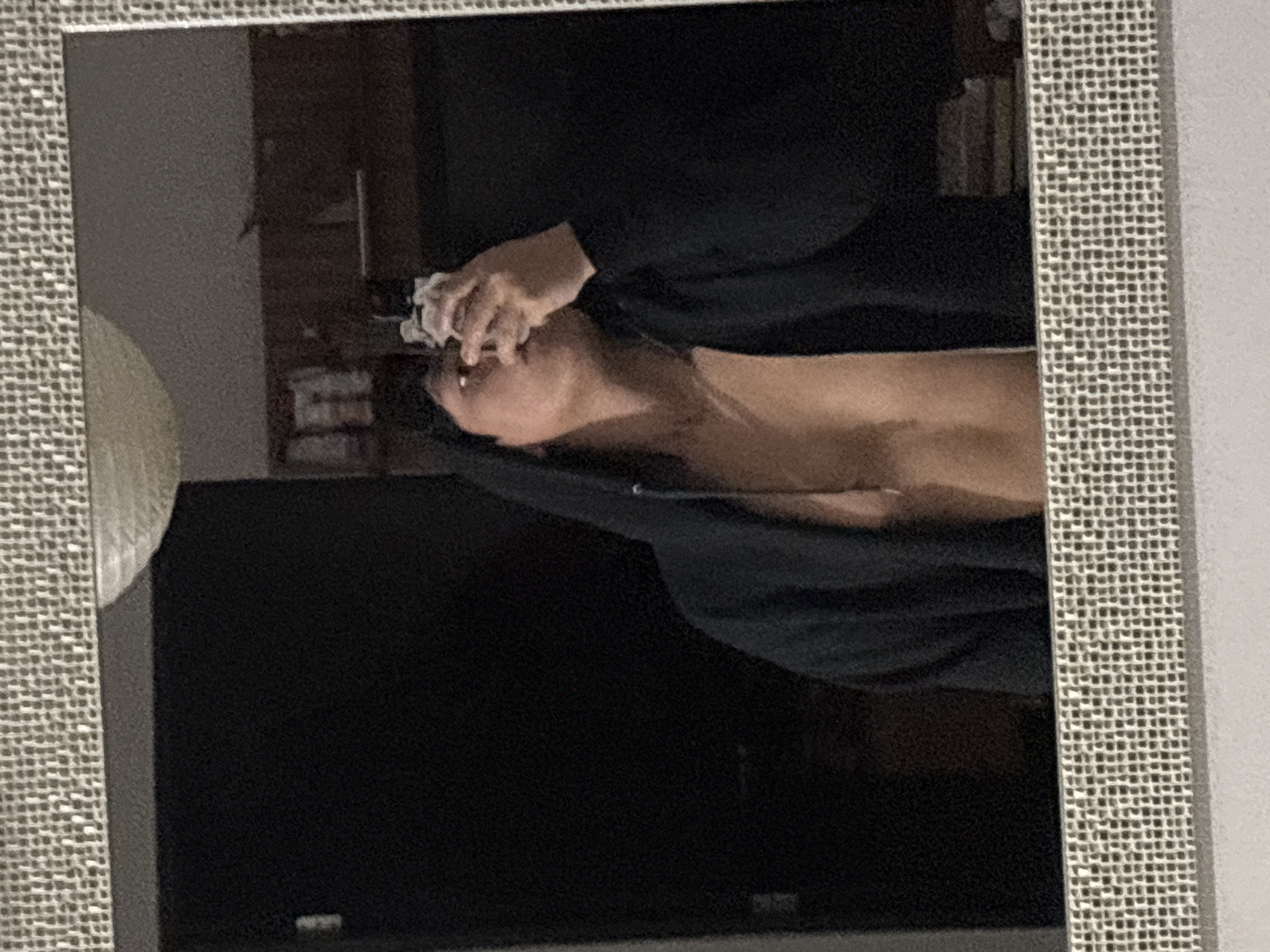 The image shows a reflection in a framed mirror. In the reflection, a person is lying down, possibly on a bed or other flat surface, wearing a black garment. The person's head is tilted back, and they are holding what appears to be a white cloth or tissue to their face with both hands. The upper part of the person's chest is visible. The background contains blurred images of furniture and other objects, indicating an interior space. A large dark rectangular object, likely a television or a large framed picture, is visible in the lower part of the reflection. The frame of the mirror is textured. There is no way to tell the location of the scene is in Mexico City.