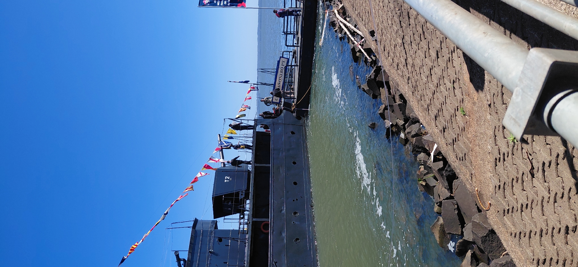 The image shows a view of the Paraguay River waterfront in Encarnación, Paraguay, on a clear, sunny day. A large, dark-colored vessel, likely a historical naval ship, is docked and decorated with a string of nautical signal flags. Several people are visible on the deck of the ship and on a smaller boat or pier adjacent to it. The water is a murky green, and the embankment is lined with dark rocks and a paved walkway. A metal railing is in the foreground, partially obscuring the view of the walkway. The ship appears to be named "CAÑONERO PY."