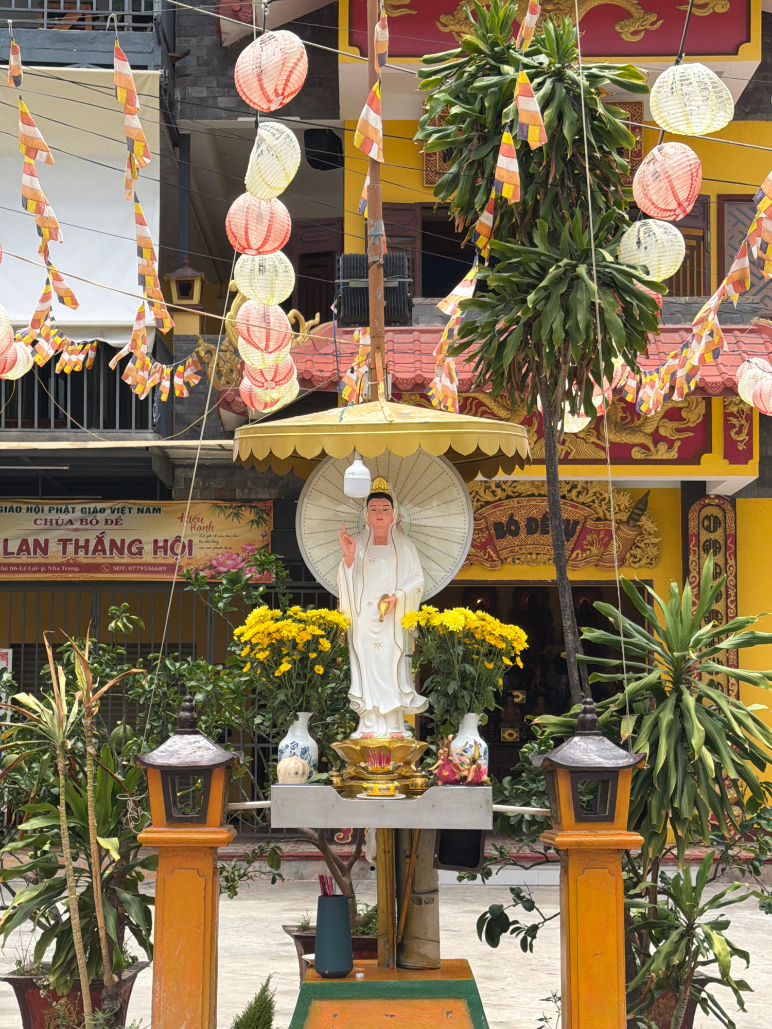 This image depicts a scene in front of a Buddhist temple in Nha Trang, Vietnam. In the foreground, a statue of a white-robed Buddha is centered, holding a golden object in its left hand and gesturing with its right. The statue is adorned with a yellow umbrella-like canopy above and sits on a golden altar surrounded by yellow chrysanthemums. To the left and right of the altar are orange lantern posts with dark roofs, and various potted plants. Behind the statue, a large white fan is visible. A banner with Vietnamese text, including "CHÙA BÔ ĐÊ" and "LAN THẮNG HỘI", hangs on a fence. The background features a yellow building with red trim and architectural details, along with strings of colorful paper lanterns and prayer flags suspended from wires. The overall setting appears to be a temple courtyard or entrance.
