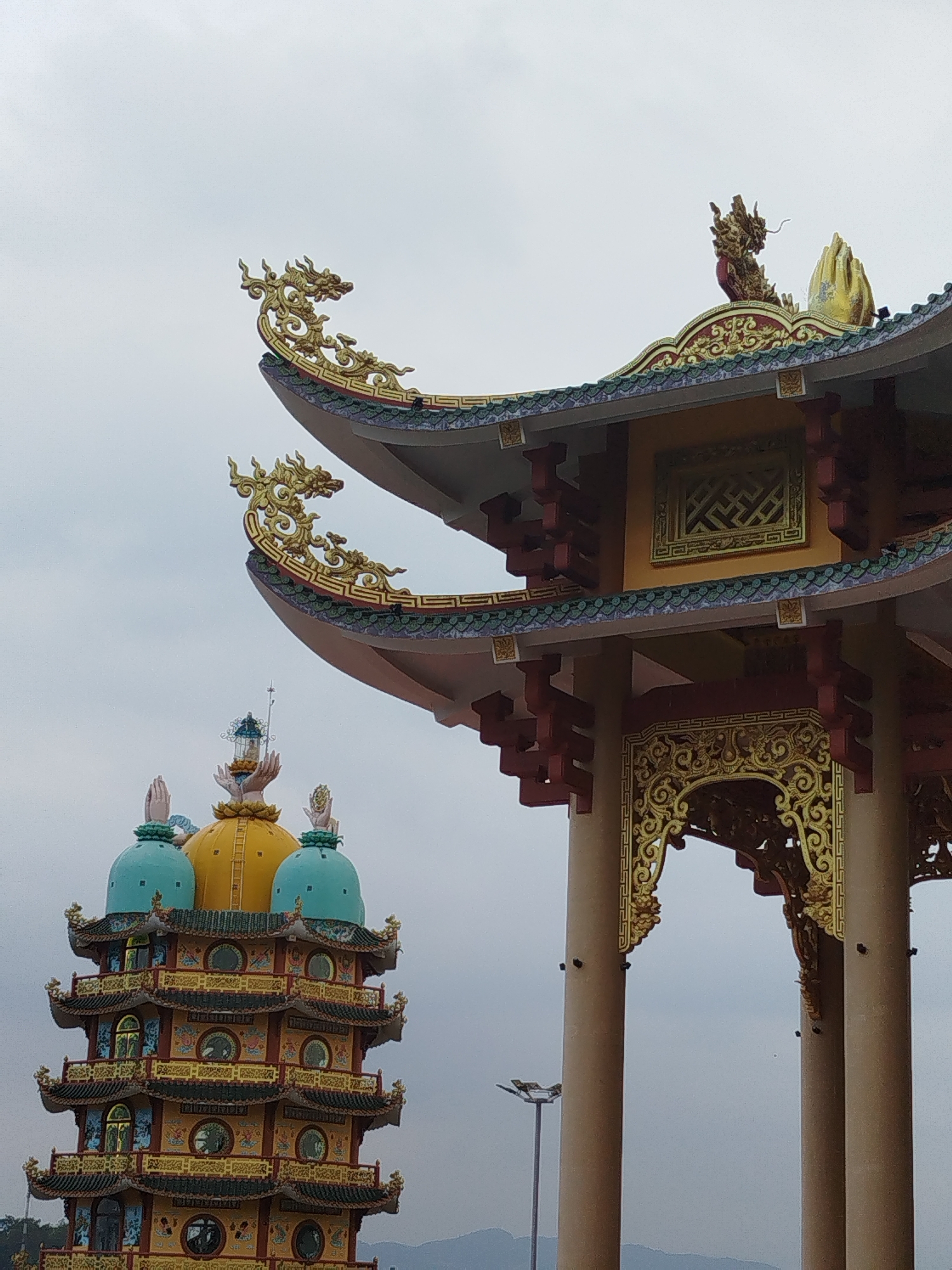 The image depicts two ornate structures in Phú An, Vietnam, against a cloudy sky. The first structure, on the left, is a multi-tiered pagoda with a yellow dome topped by hands holding a lotus. It has green domes on either side. The other structure, on the right, is a gate-like structure with red and gold accents. Dragon statues adorn the roof corners of the gate. The pillars supporting the gate are beige. A streetlight is visible in the background, near the base of the gate structure.