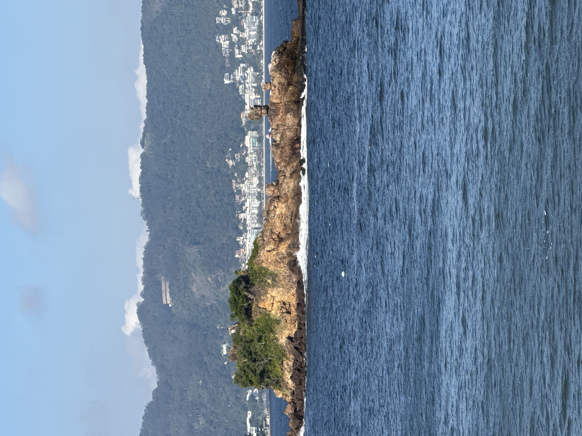 This is a photograph of a rocky island covered in dense green vegetation, situated in the ocean. The island is brown and orange in color and has rough textures. Waves are crashing against the shore of the island. In the background, a city with white buildings and a lush green mountain range can be seen. The sky is blue with some white clouds. The image appears to have been taken from a boat or a coastal location. This location is likely the coast of Niterói, Brazil, which is known for its islands, beaches, and mountainous landscape.