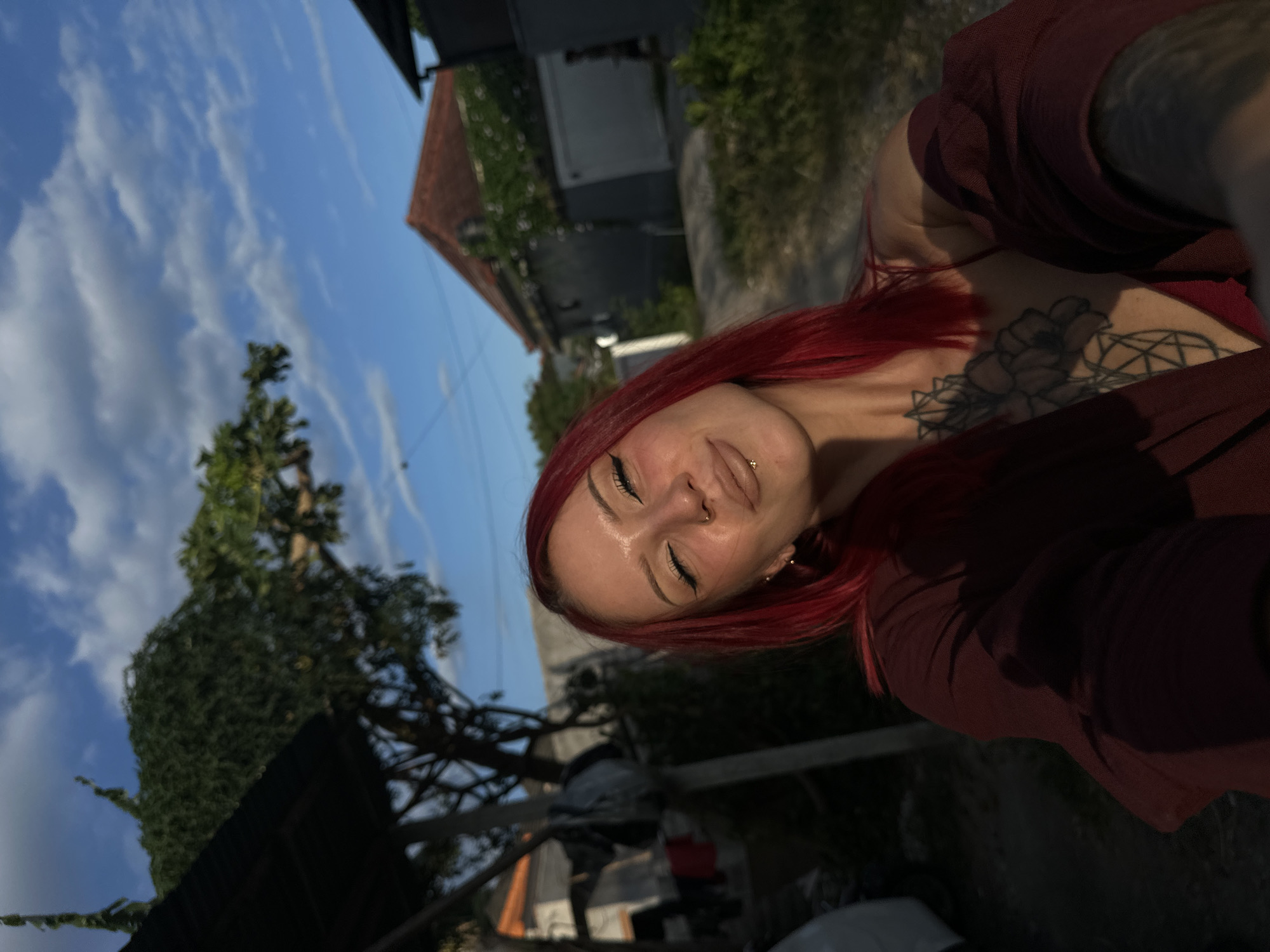 A selfie shows a young woman with fair skin and bright red hair. She has her eyes closed and wears several piercings. She is wearing a dark red, loose-fitting top and has a tattoo on her upper chest. The background includes a bright blue sky with wispy clouds, a building with a dark gray exterior and a red tile roof, and lush greenery. In the lower left, there's a glimpse of a structure with a dark, possibly corrugated roof and some obscured objects underneath. Based on the general environment, the photo could be from North Kuta, Indonesia.