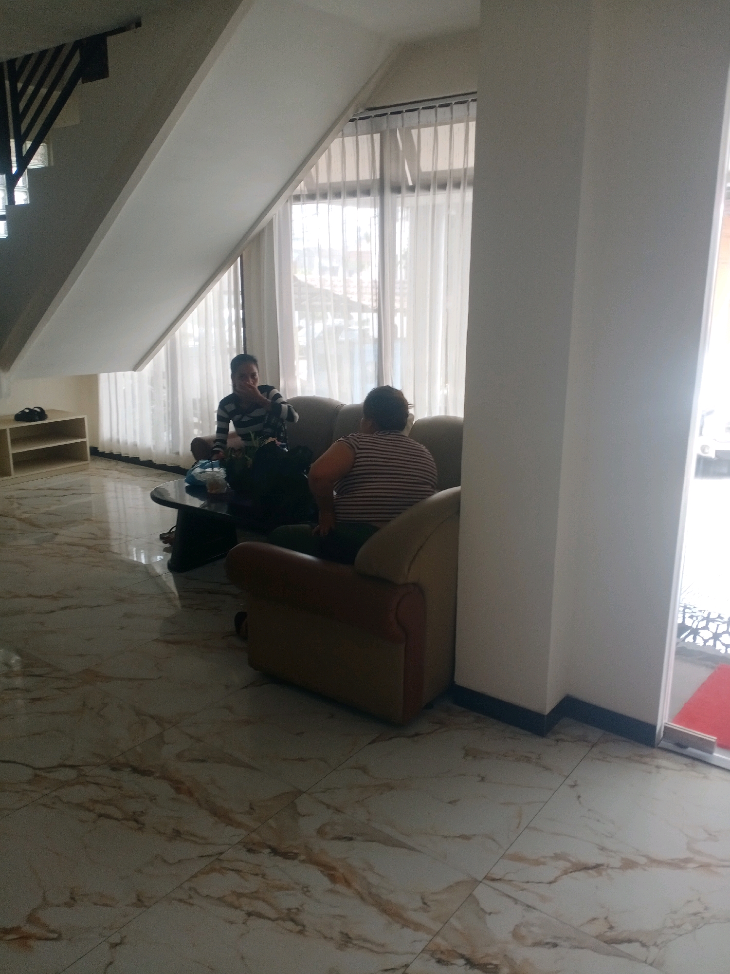 The image depicts an indoor scene, possibly a hotel lobby or lounge, with marble flooring. Two individuals are seated on a couch. The person on the left is wearing a striped shirt and appears to be reaching towards the table in front of them. The person on the right, also in a striped shirt, is seated with their back to the camera. A coffee table with items on it, including bags and papers, is positioned in front of the couch. Behind the couch, sheer white curtains cover a large window. To the left of the couch, a staircase with a slanted white ceiling above it is visible, along with a white shelf unit. The overall ambiance suggests a relaxed, indoor setting. Based on the available information, this location is identified as being in Samarinda, Indonesia.