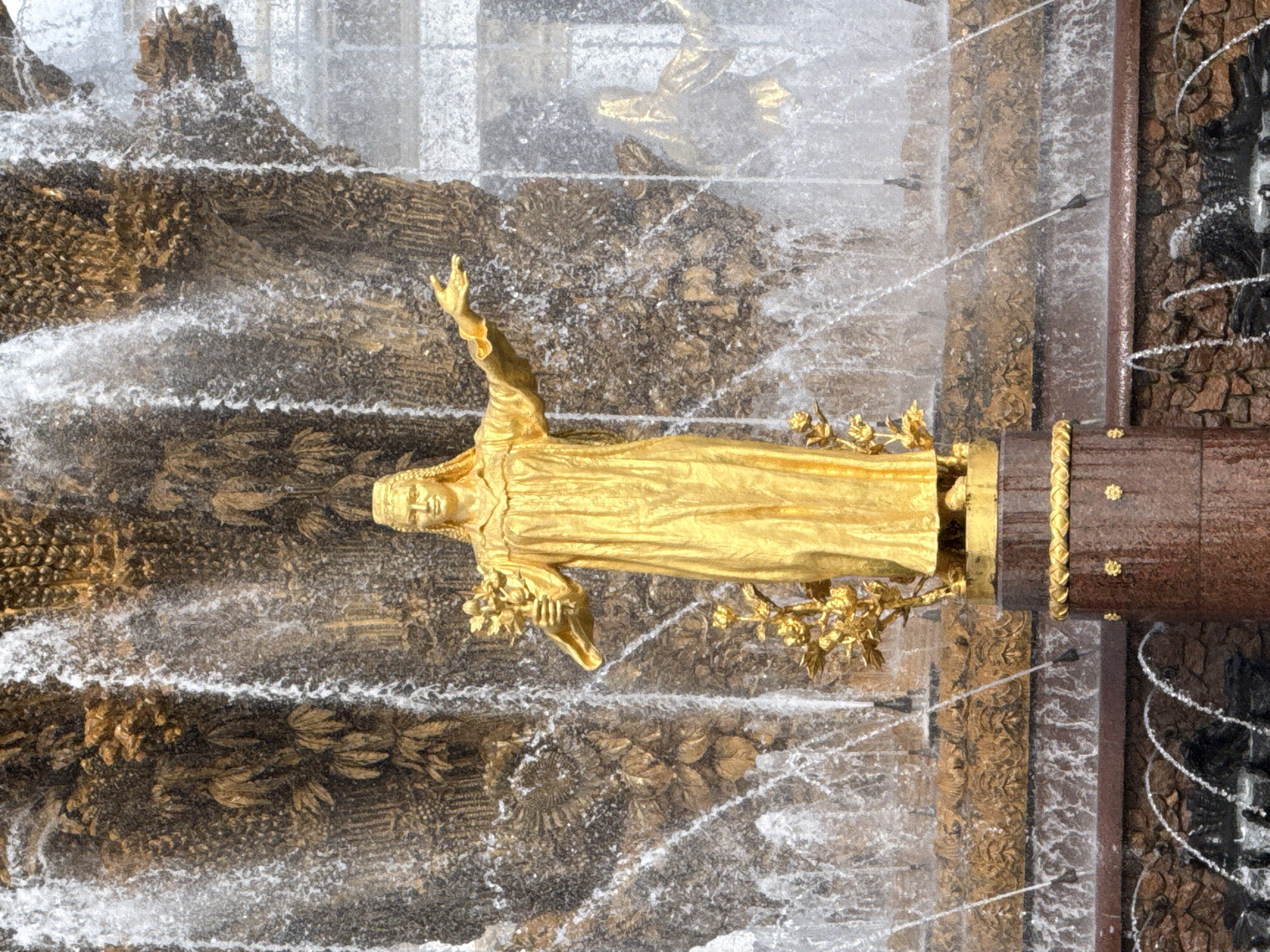 The image shows a golden statue of a woman holding a bouquet of flowers in front of a large, multi-tiered fountain. The statue is detailed, with the woman wearing a long, flowing gown and a headdress. Water cascades down the stone and golden decorative elements of the fountain, creating a dynamic and opulent backdrop. In the background, a distorted reflection of another golden statue can be seen through the falling water. The fountain itself appears to be made of stone and metal, with intricate carvings and decorative details. The overall impression is one of grandeur and artistry. This is the "Friendship of Peoples" fountain located in VDNKh Park in Moscow, Russia.