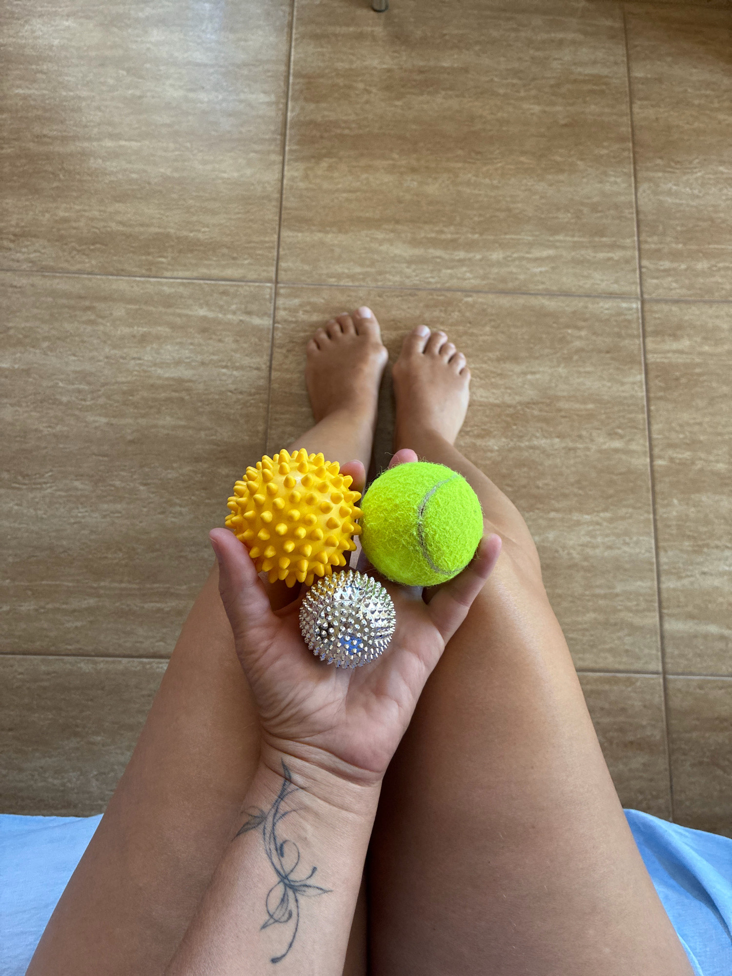 An overhead shot shows a person's legs and feet on a tiled floor. The person is holding three balls in their hand: a yellow spiky ball, a silver spiky ball with blue accents, and a green tennis ball. The person has a tattoo on their left forearm. The floor tiles are a light brown color. This scene is taking place in the city of Sochi, Russia.