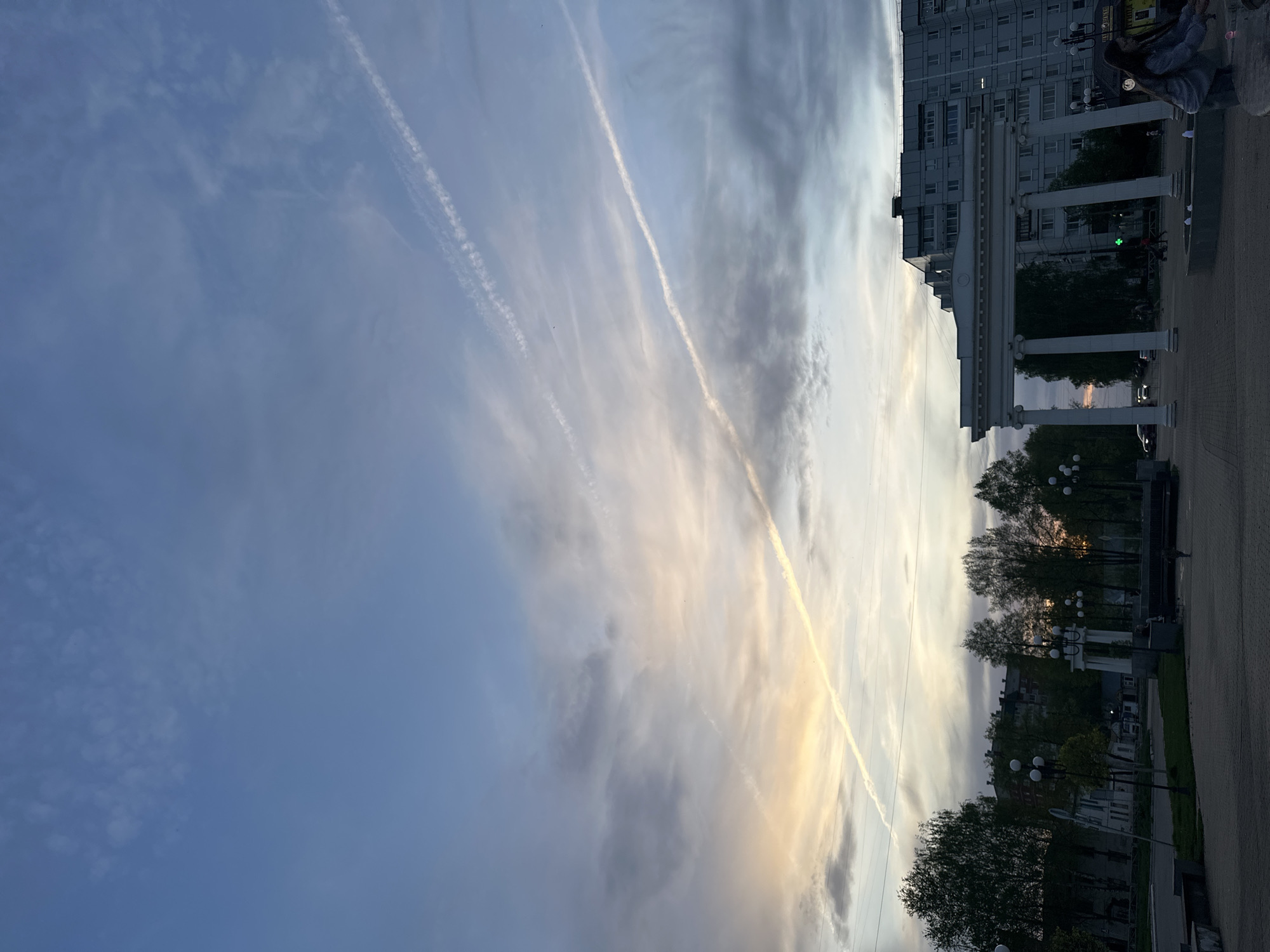 The image shows a view in the city of Chaikovsky, Russia, likely taken in the evening based on the lighting. The sky is streaked with clouds and contrails from aircraft. Part of a building with white columns is visible on the right side of the frame. Below the building, there are trees and street lamps, suggesting a park or street scene. A person wearing a hooded shirt is visible in the upper right of the image.