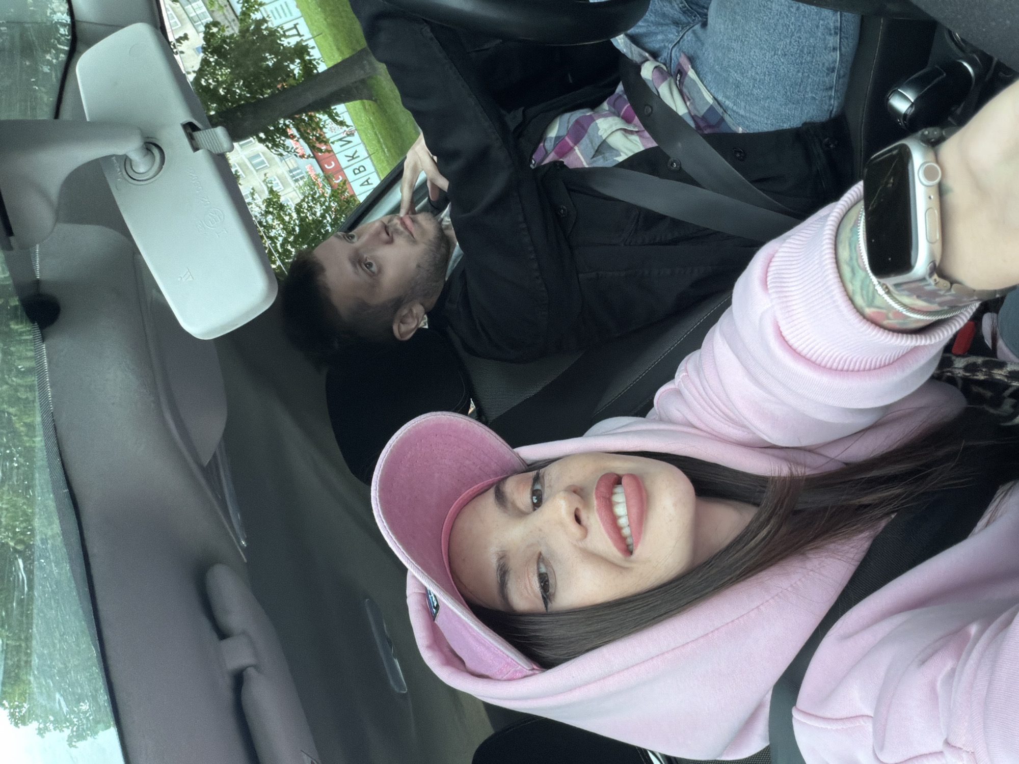 A slightly rotated interior shot from a motor vehicle shows two people, a fair-skinned woman and a man, both wearing seatbelts. The woman is closer to the camera. She wears a pink hooded sweatshirt and a matching baseball cap. The man is in the background, wearing a dark jacket over a plaid shirt. The vehicle is parked, as visible through the windshield is a reflection of an urban scene, including a building with signage in Russian, which suggests the location is Saint Petersburg, Russia. A rear-view mirror is visible in the upper left corner of the frame. The woman's left wrist is prominent, showing a silver smartwatch with a metal band. The overall lighting is bright, suggesting daytime.