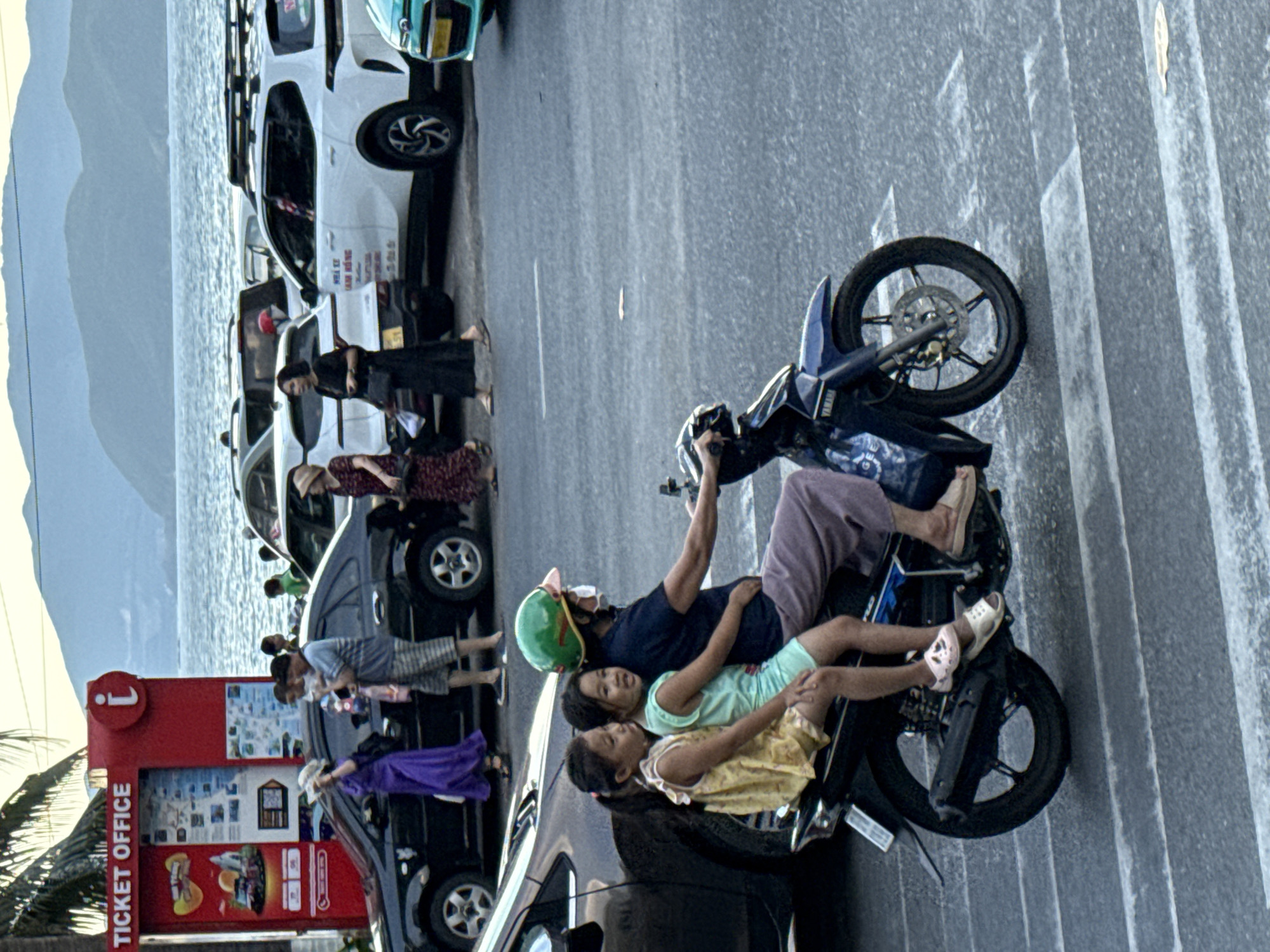 A family of four is riding on a motorcycle on a street, with a body of water and mountains in the background. The father, wearing a dark blue t-shirt and purple pants, is driving. His two daughters are passengers. The older daughter, wearing a light green shirt and yellow skirt, is sitting between her father and younger sister. The younger daughter, wearing a yellow dress, is sitting in front of her father. The family appears to be enjoying a sunny day, possibly in the late afternoon given the long shadows. The scene is likely in Nha Trang Ward, Vietnam, as suggested by the location context, with the sea visible to the left and mountainous terrain in the distance.
A red ticket office structure with the "i" logo and "TICKET OFFICE" text is visible on the left side of the image. It displays various maps and information. Several parked cars are also present, including a white SUV and a black car. The overall atmosphere seems relaxed and everyday, depicting a common mode of transport and family activity in the region.