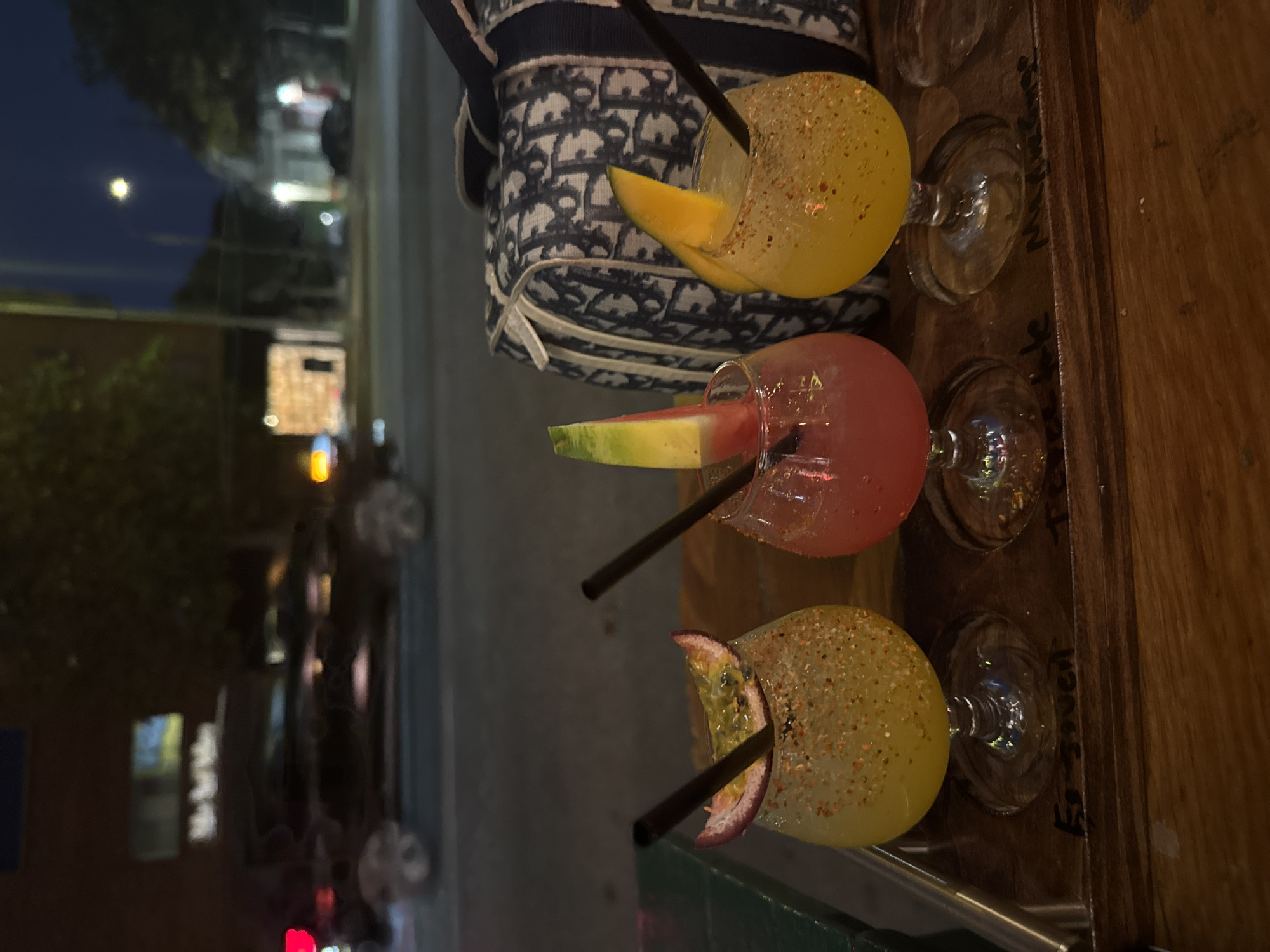 The image shows three cocktails, each with a black straw and fruit garnish, lined up on a wooden surface. The topmost cocktail has a yellow drink and a mango garnish. The middle cocktail is pink with a watermelon garnish. The bottom cocktail has a yellow drink with a passion fruit garnish. Each of the cocktails are sitting in a glass. In the background, there is a blurred scene of a street at night, with city lights. This scene suggests a location in a city, likely Los Angeles, United States of America. There are also some cars parked on the street.
