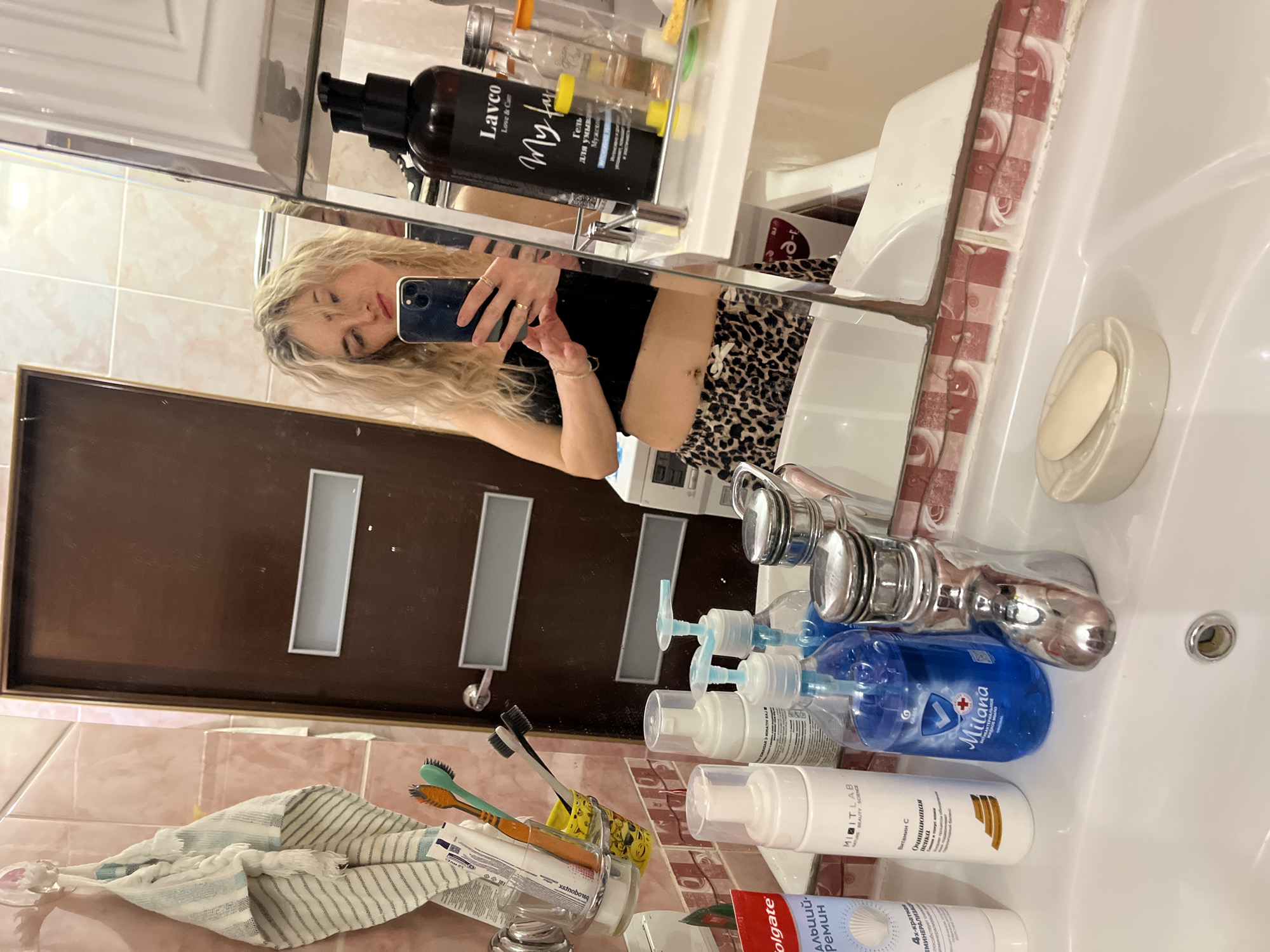 The image shows a bathroom mirror selfie of a woman with blonde, curly hair. She is holding a phone in her right hand, capturing her reflection. She is wearing a black top and leopard-print bottoms. The bathroom setting includes a sink with various toiletries and personal care products. There are toothpaste, toothbrushes, and bottles of soap and lotion. The mirror reflects the door and adjacent wall, revealing a hint of other items in the bathroom, such as a towel. The branding on one of the soap bottles ("Milana") is the key to knowing this is Chaikovsky, Russia.