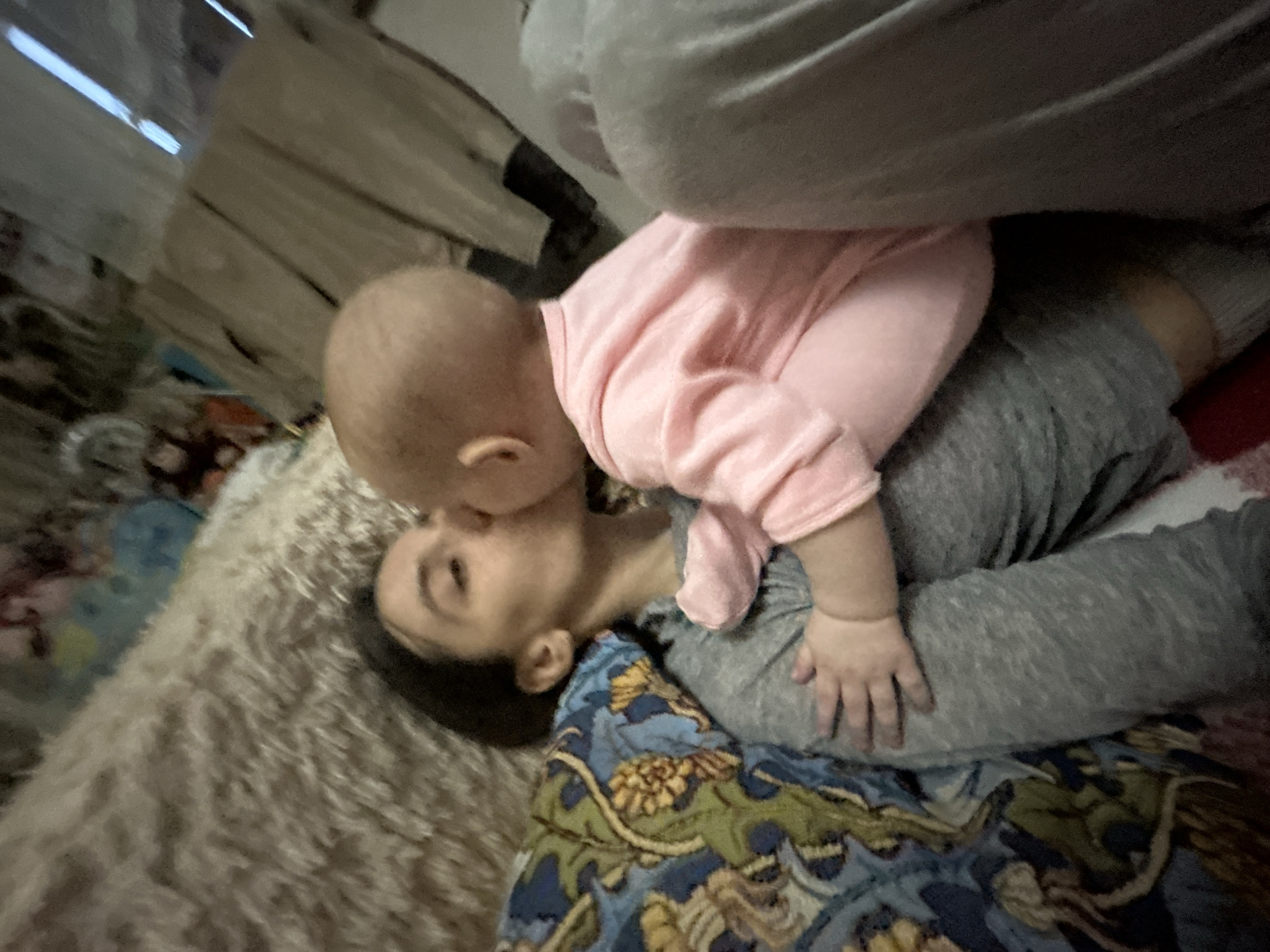 A baby in a pink shirt is lying on top of an adult wearing gray clothing and lying on a patterned blanket. The adult's head is visible, with dark hair. The blanket has a blue and yellow floral pattern. A fuzzy, tan rug is visible to the left of the blanket. The background is blurred and shows a wall or other structure with light-colored wooden slats. There is no discernible information to determine the location is in the city of Moscow, Russia.