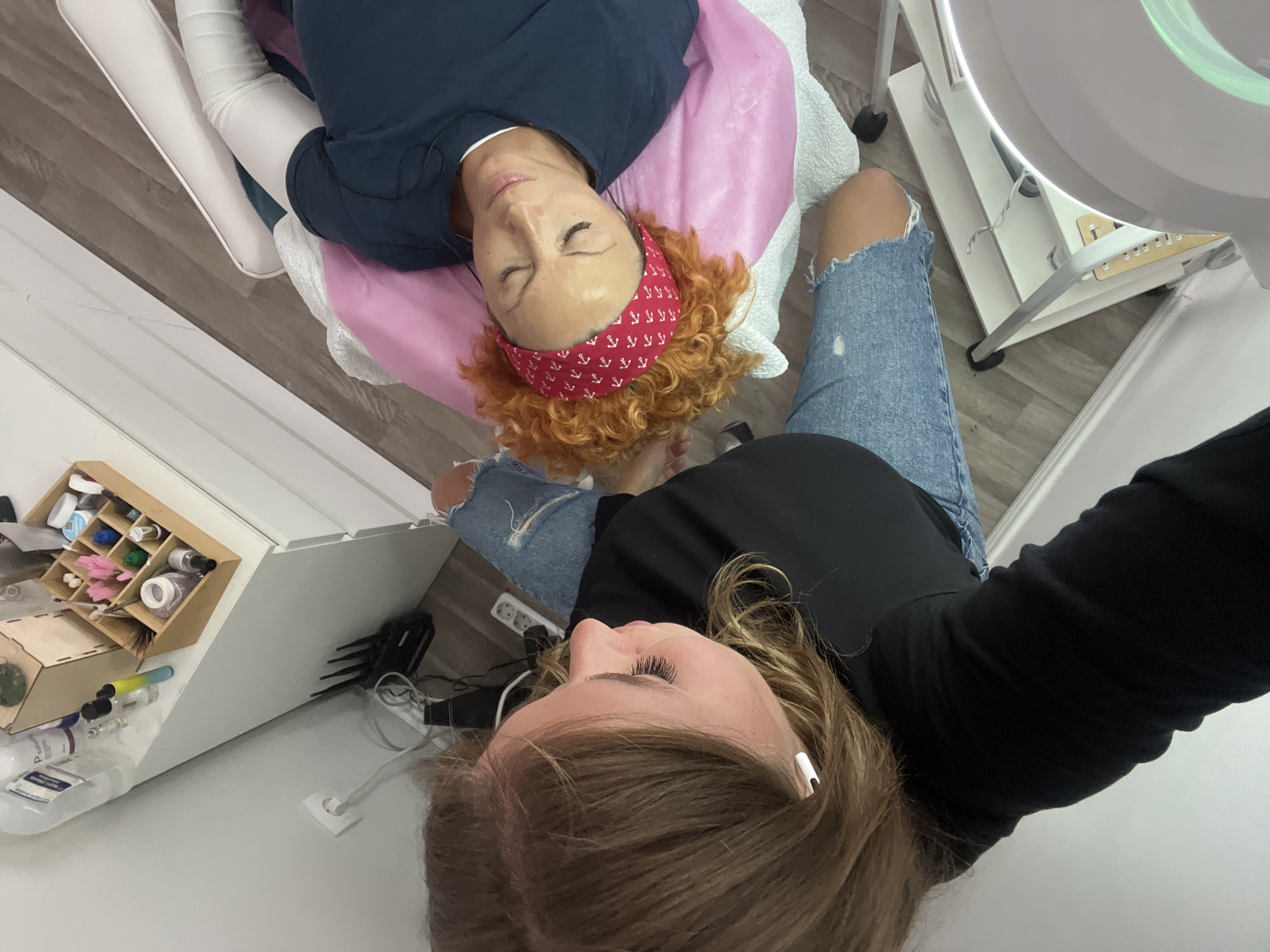 This high-angle image captures an indoor scene within what appears to be a beauty salon or treatment room. Two women are the primary subjects.
In the foreground, a woman with long, light brown hair wears a black long-sleeved top and ripped blue jeans. A white earbud is visible in her right ear, and she has pronounced eyelashes. She appears to be the one taking the photo, looking down towards the other individual.
Lying on her back on a white treatment table, with her head on a pink pillow, is the second woman, likely a client. She has distinctive reddish-orange curly hair, contained by a red bandana featuring white anchor patterns. She is wearing a dark blue top, possibly scrubs, and her eyes are closed, indicating a state of relaxation or being amidst a procedure.
The setting includes a light wooden plank floor and white cabinetry. To the right of the client, a large, white circular lamp on a stand, commonly used for aesthetic treatments, is visible. In the bottom left, a white counter holds a wooden organizer filled with various beauty supplies such as small bottles and cotton-like items. A white power outlet is also seen on the wall/floor.
The activity depicted is a beauty treatment, likely involving the client's face or eyelashes, being performed by the woman in the foreground. The overall atmosphere is professional and focused. This scene is located in Budva, Montenegro.