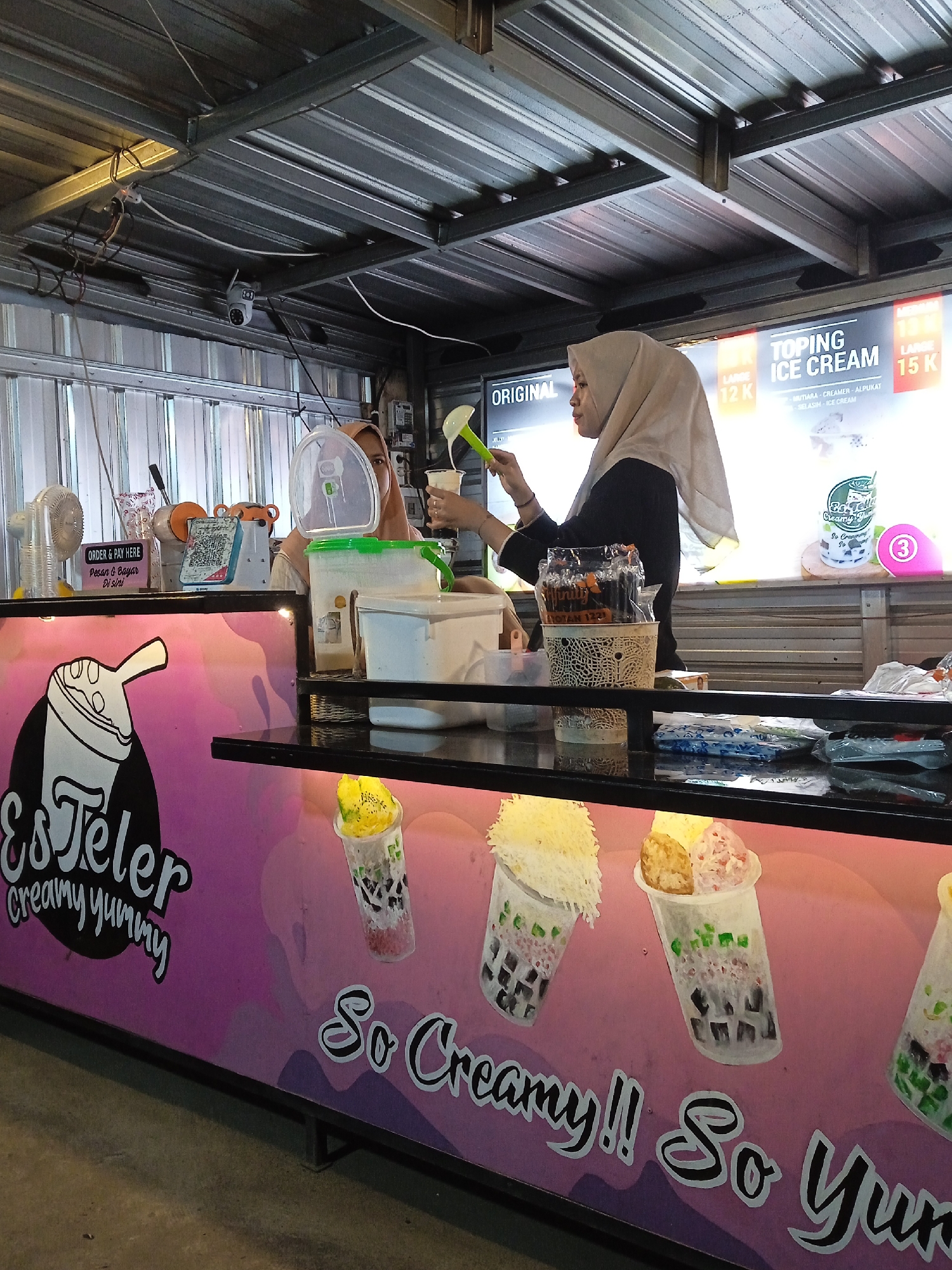 An indoor or semi-open food stall, likely in Sekopek Wetan, Indonesia, is depicted, specializing in a creamy dessert called "Es Teler." Two women are working behind a counter with a black top and a vibrant pink and purple branded front. The woman on the right, wearing a light-colored hijab and a black long-sleeved shirt, is actively preparing a drink or dessert, scooping ingredients from a large white container into a clear plastic cup. Another woman, also in a hijab, is partially visible behind other containers on the left side of the counter.
The stall's branding "Es Teler creamy yummy" is prominent on the counter, along with the slogan "So Creamy!! So Yummy" (partially visible). Illustrated cups filled with colorful desserts are also part of the counter design. Above the counter, menu boards display prices and options: "ORIGINAL 12 K" (Rupiah), "TOPING ICE CREAM LARGE 15 K", and topping choices like "MUTIARA - CREAMER - ALPUKAT", "SELASIM - ICE CREAM". A small sign on the left reads "ORDER & PAY HERE" and "Pesan & Bayar Di sini". A white fan is placed on the left side of the counter, and a security camera is mounted on the corrugated metal ceiling structure, which also has exposed wiring. A container on the counter has the text "STAN 1221". The overall setting suggests a casual food preparation area, possibly within a market or a small eatery.