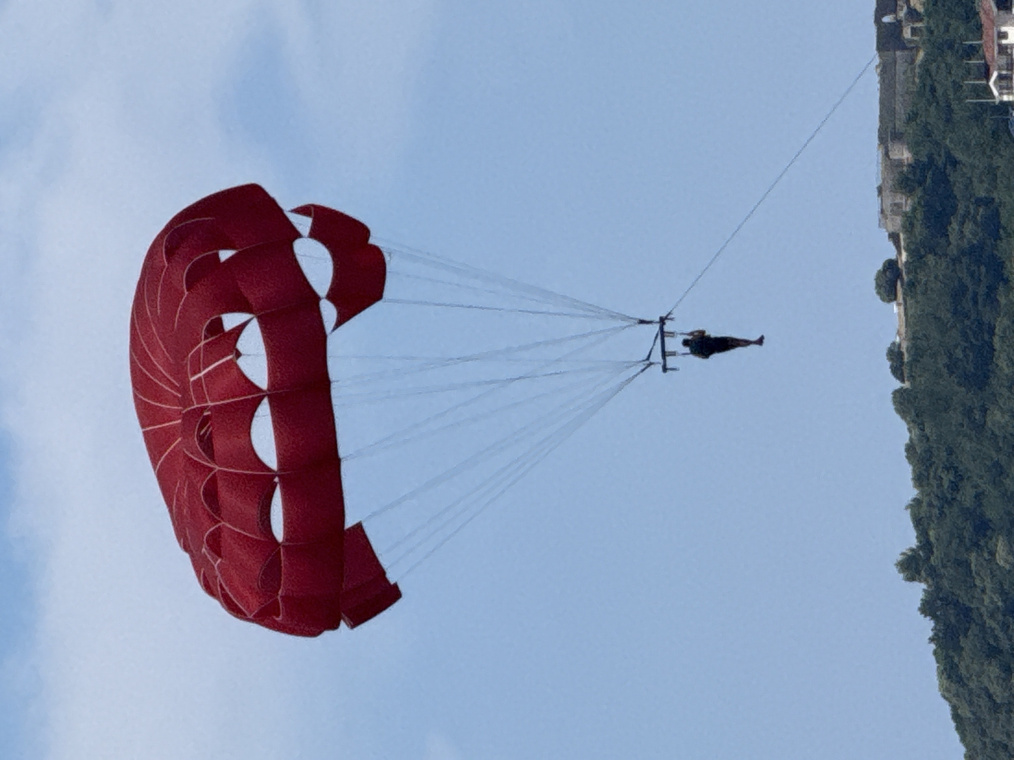 An individual is parasailing high in the sky above a lush, green hillside and a historic stone structure, possibly a fortress, in the city of Budva, Montenegro. The parasail, a vibrant red, dominates the upper portion of the frame, its fabric taut against a clear blue sky dotted with faint clouds. Below the parasail, thin white cords connect to a person suspended in mid-air, their dark silhouette stark against the bright sky. The person appears to be facing away from the viewer. The hillside below is densely covered with trees, creating a textured green expanse. A portion of an old stone building with arched openings is visible on top of the hill, suggesting a historical site. The overall scene captures a moment of recreational activity against a backdrop of natural beauty and historical architecture.