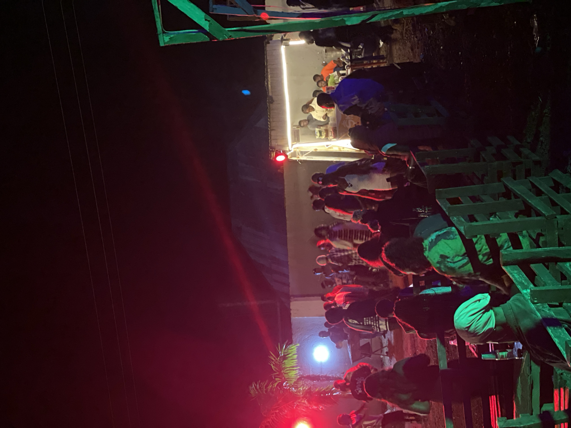 The image depicts an outdoor nighttime gathering in Durumi, Nigeria. A group of people are visible, some standing and socializing, while others are seated on wooden benches. Red and green lights illuminate the scene, suggesting a festive or event atmosphere. In the background, a food truck or service counter is set up, with individuals appearing to be serving or interacting with customers. A palm tree is also visible to the left. The ground appears to be dirt or rough terrain. The overall impression is a lively community event taking place after dark.
