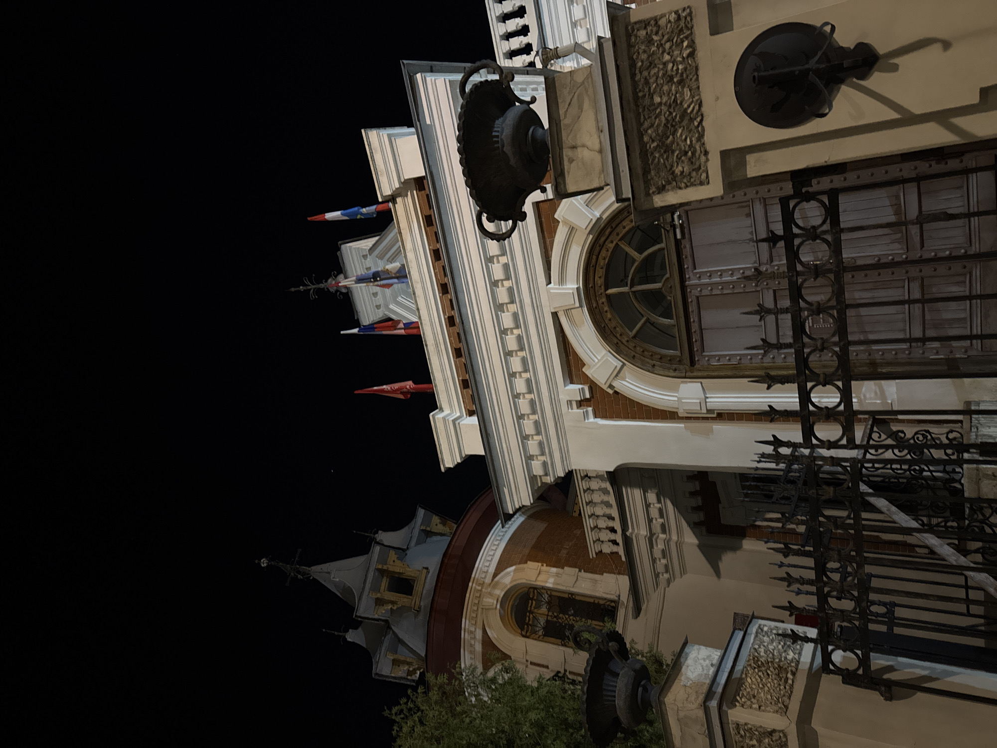 The image shows a building at night. The building is brightly lit, with multiple windows and architectural details visible. At least two flags, one red and the other with a red and blue design, are displayed. A decorative lamp is attached to the building's exterior. A wrought iron fence is visible in the foreground, and a small amount of greenery can be seen at the bottom. The rest of the image is a black sky.