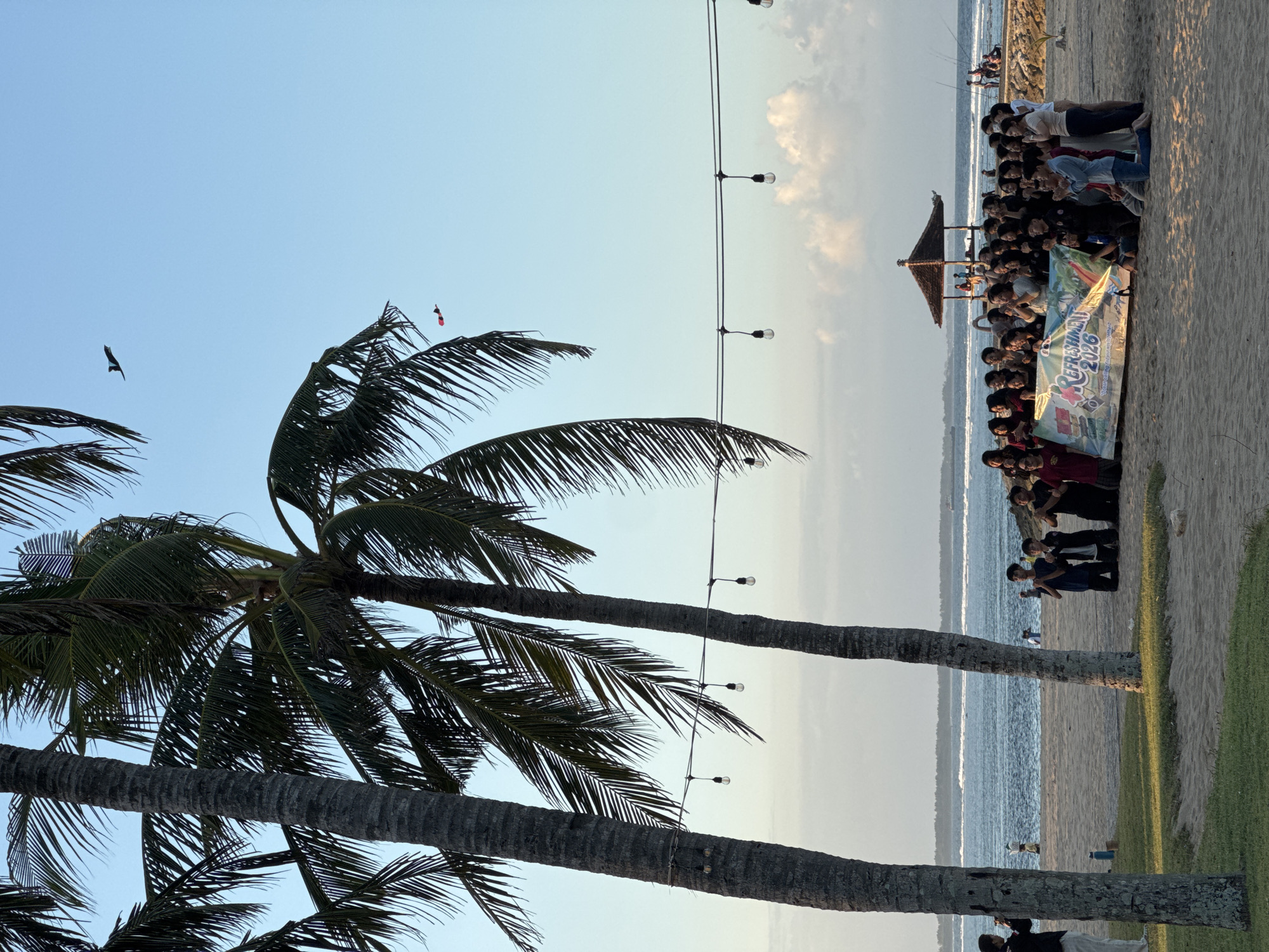 In Kuta Selatan, Indonesia, a group of young people, dressed in various casual attire, are gathered on a sandy beach, posing for a photograph with a banner that reads "REFRESHMENT 2026." The banner, featuring colorful graphics and text, is held horizontally across the front of the group. Some individuals are kneeling, while others are standing or leaning, all facing towards the camera. Many are clapping their hands, suggesting an enthusiastic atmosphere.
Behind the group, the ocean stretches out to the horizon, with gentle waves lapping the shore. A breakwater, made of rocks, extends into the sea, with a small thatched-roof pavilion situated on it. A few distant figures can be seen on the breakwater and in the water. The sky above is a clear, pale blue, with a few scattered clouds. Two kites are visible in the sky, one a dark silhouette and the other a red and black object.
In the foreground, two prominent palm trees with rough bark and large fronds frame the left side of the image, their leaves casting shadows on the sand. String lights with exposed bulbs are strung between the trees, adding a decorative element to the scene. To the far left, a person in white pants and a dark top is seen from the back, looking towards the ocean. The beach has areas of sand and patches of green grass. The overall impression is a casual, celebratory gathering at a tropical beach location.