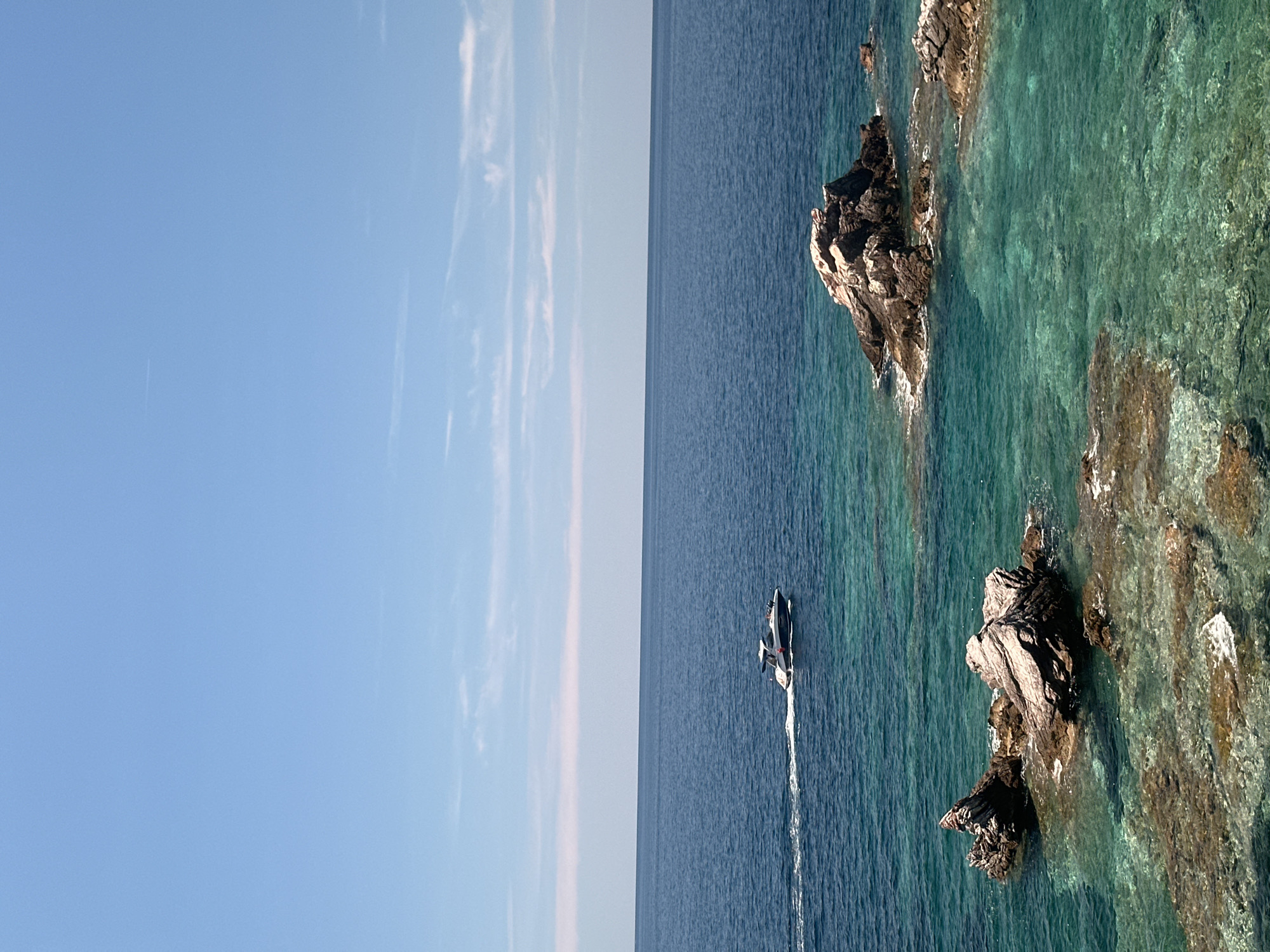 A scenic view of the Adriatic Sea is captured from the coast near Sveti Stefan, Montenegro. The image showcases the vibrant turquoise waters near the shore, with several rocky outcrops breaking the surface. These rocks, varying in size and texture, are partly submerged and appear weathered by the sea. In the mid-ground, a white motorboat with a dark hull cruises through the deeper blue waters, leaving a white wake behind it. The horizon is a sharp line where the deep blue sea meets a clear sky, adorned with a few wispy white clouds. The overall impression is one of a serene and beautiful coastal landscape under a bright, sunny day.