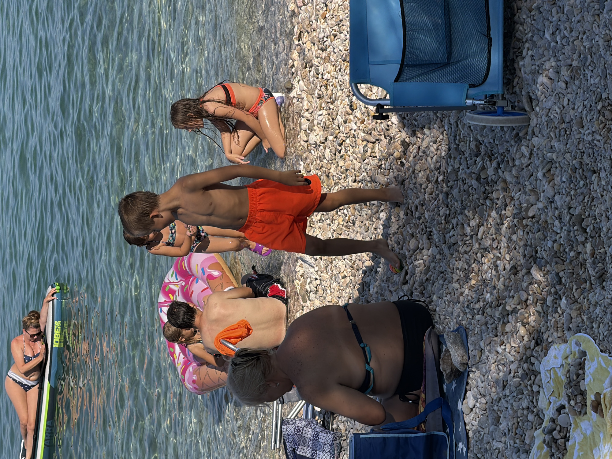 This media file depicts a sunny day scene at a pebble beach in Općina Baška, Croatia. Several people are enjoying the water and shoreline. In the water, a woman in a polka dot bikini is holding onto a paddleboard. Nearby, a young boy in orange shorts is standing in the shallow water, with another child partially visible in the background. A pink and white inflatable ring is floating in the water, with a couple embracing on it. On the beach, a young girl in an orange bikini is kneeling. Further back, a person is sitting in a blue beach chair, with another person partially visible in a black swimsuit lying down. The sun appears to be high in the sky, suggesting it is midday, and the weather seems warm and clear. The beach is covered in smooth, grey and white pebbles, and the water is clear and blue. A blue wheeled cart is visible on the beach. Some towels are spread out on the pebbles. Text is visible on the paddleboard: "10BE" and "JARRA".