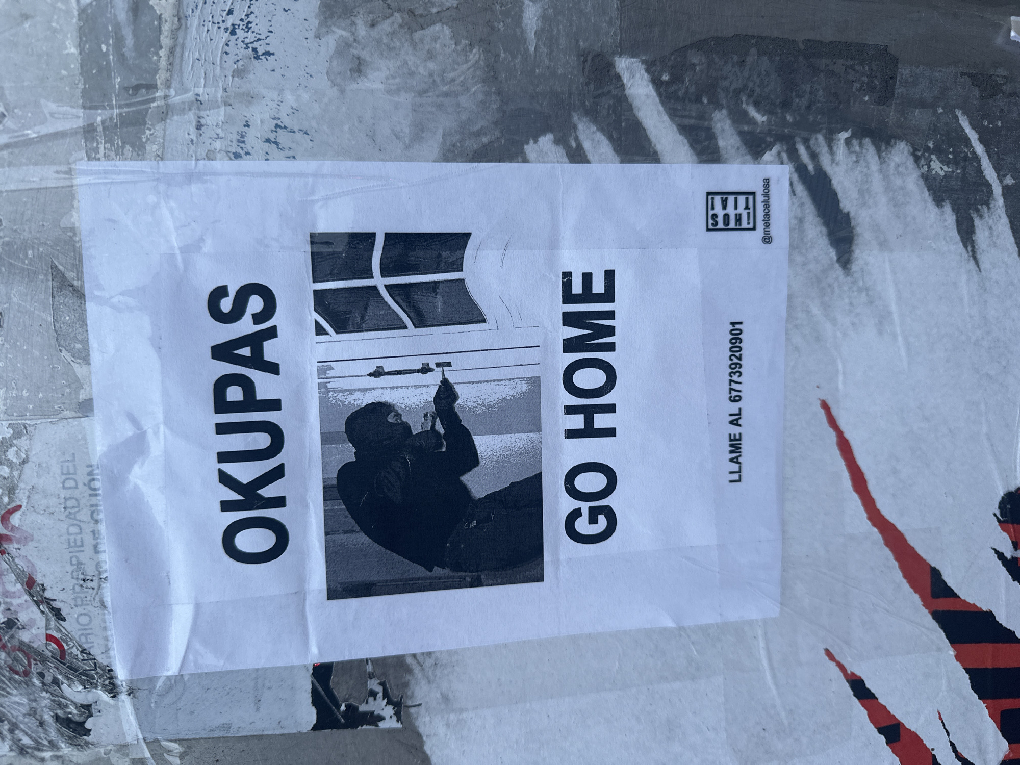A white paper poster is affixed to a textured, weathered wall or surface in Gijón, Spain, confirmed by visible text fragments "PROPIEDAD DEL ... DE GIJÓN" on an underlying poster. The poster displays the capitalized word "OKUPAS" at the top. Below this, a black and white image depicts a person, clad in dark clothing and a balaclava, actively prying open a white door with a tool. The door features a glass-paneled window. Beneath the image, the capitalized phrase "GO HOME" is printed. At the bottom of the poster, contact information reads "LLAME AL 6773920901," followed by a square black logo with vertical text "¡HOS TIA!" and "@metacelulosa." The poster exhibits minor wrinkles and is partially covered by a translucent adhesive layer. The background surface is a mix of faded grey and white, showing remnants of other ripped posters, including red and black striped fragments on the lower left.