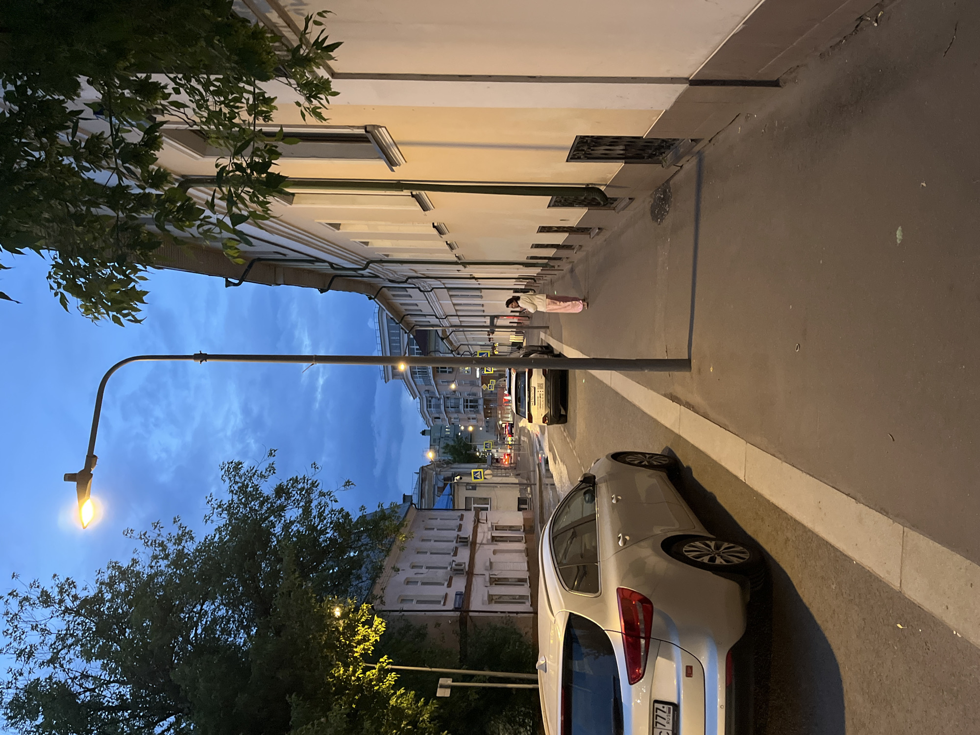 The image depicts a street scene in an urban environment, possibly at dusk or early evening. The sky is a deep blue with some clouds visible. A street lamp is illuminated, providing light to the immediate vicinity and the adjacent tree foliage. A row of buildings lines one side of the street, their facades a light cream color. A person is standing on the sidewalk, dressed in light-colored clothing, possibly reading or looking at a phone. A white car is parked on the right side of the street, partially obstructing the view. The asphalt road surface is dark, and the overall atmosphere is calm and still. The location is Unknown, Unknown.