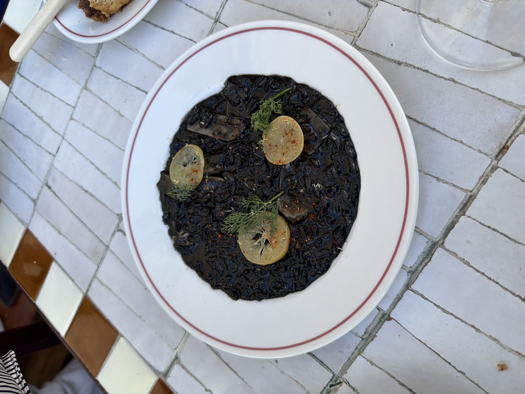 A top-down, medium shot captures a white plate with a red rim filled with squid ink pasta. The pasta is a deep black, glistening and mixed with what appears to be seafood. Three thin slices of lemon, sprinkled with red pepper flakes, are placed on top of the pasta. A sprig of green dill garnishes the dish. The plate rests on a white tiled surface with grey grout lines, arranged in a brick pattern. In the background, the edge of a table with brown and cream colored tiles is visible, along with a portion of a person's black and white striped sleeve. To the right, the rim of a wine glass is visible, and a small portion of another dish with a piece of pastry can be seen. The setting suggests an outdoor dining area, possibly a restaurant in London, United Kingdom.