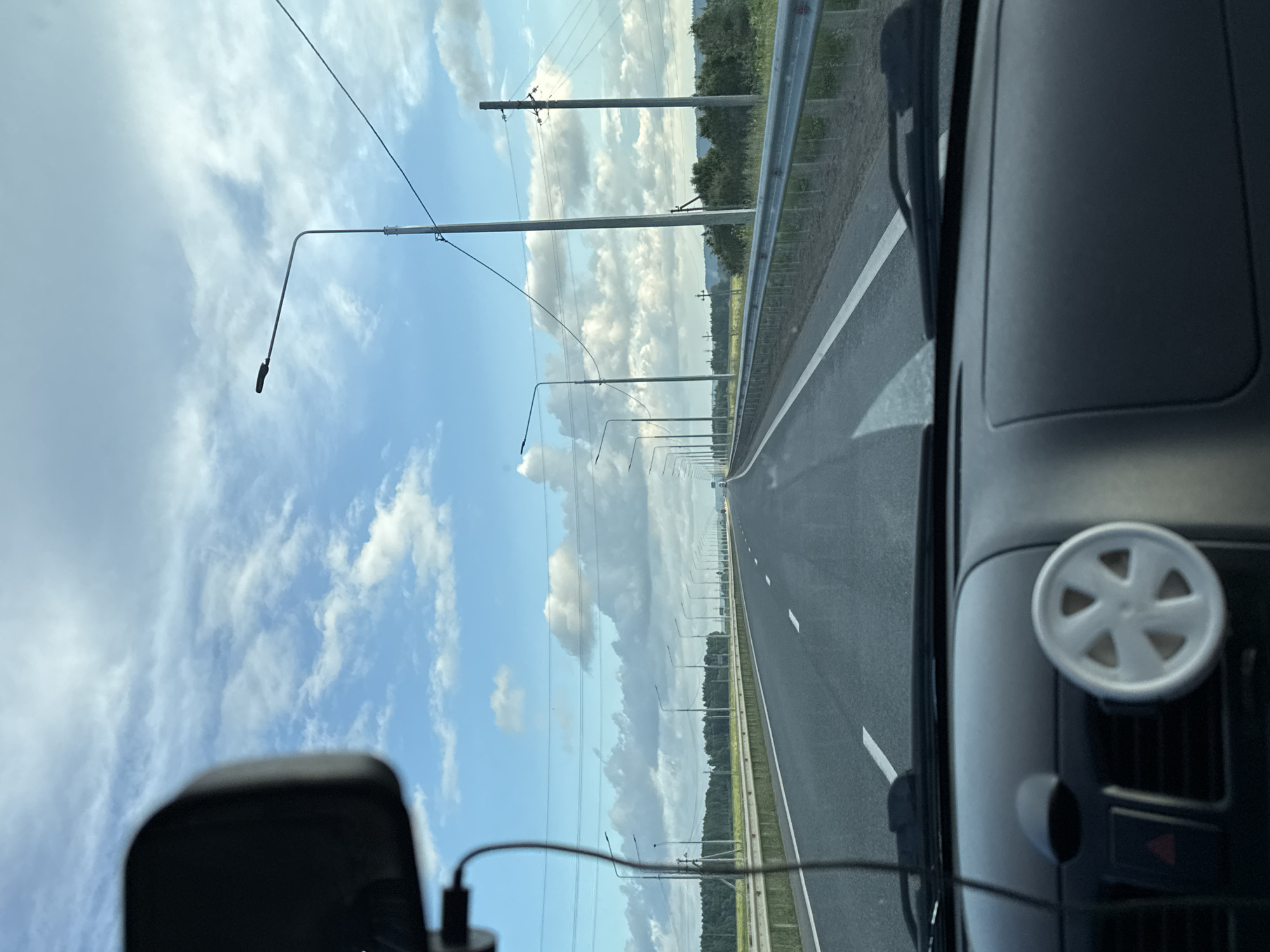 The image is taken from inside a vehicle, looking out through the windshield at a highway in Krasnokamsk, Russia. The road stretches into the distance, with multiple lanes marked by white lines. On the right side of the road, there is a metal guardrail, and beyond that, a grassy verge and trees. Streetlights line the highway on both sides, spaced at regular intervals. The sky is partly cloudy with fluffy white clouds against a blue background. The dashboard of the car is visible in the foreground, with a small, white, six-spoke decorative item attached to the air vent. A black rearview mirror is also visible in the upper left corner of the frame. The scene appears to be captured during daylight hours, with good visibility. There are no people or other vehicles immediately visible on the road. The overall impression is of a typical highway scene during a drive.