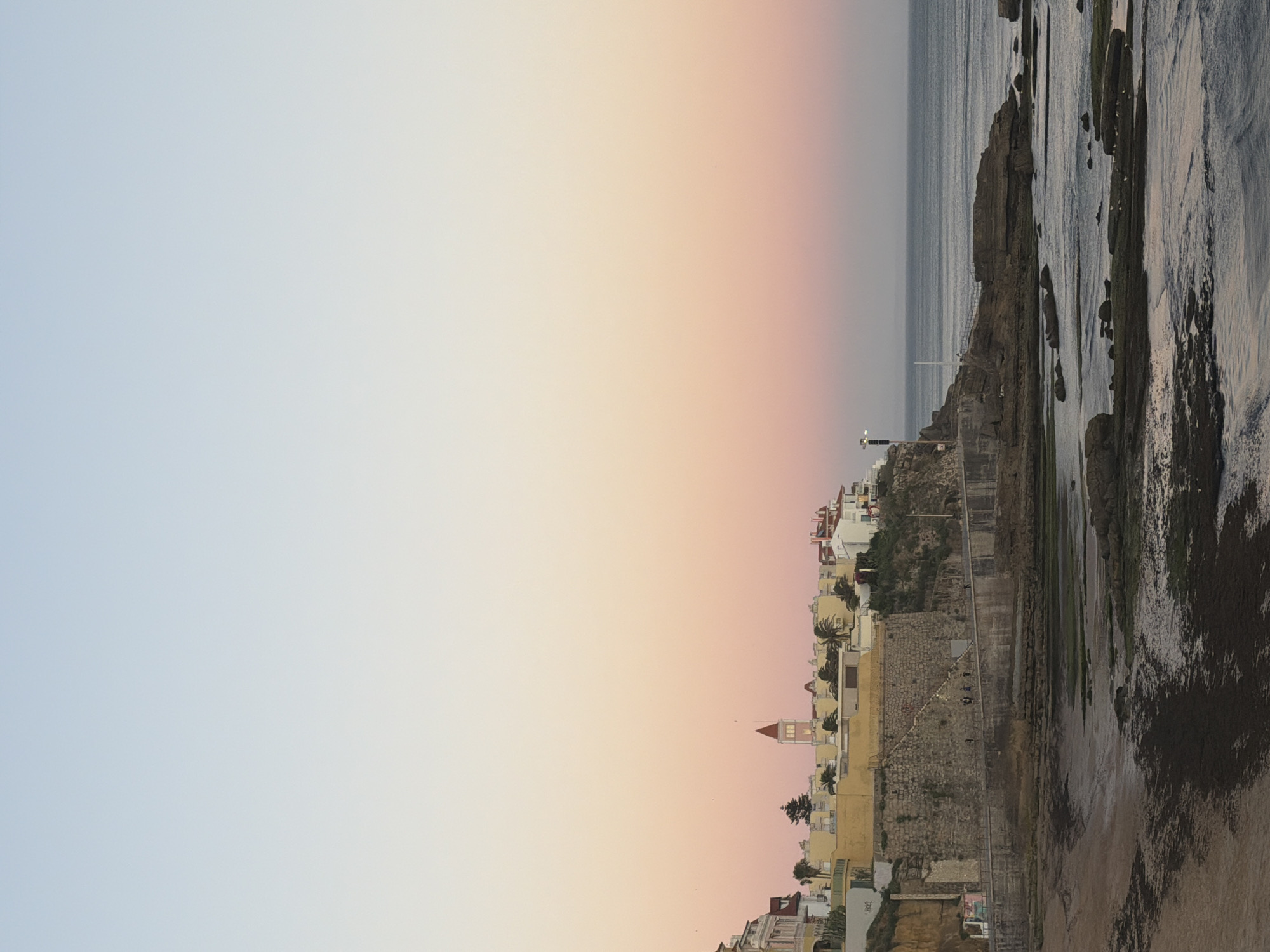 The image depicts a panoramic view of the Cascais coastline in Portugal during a sunset. The sky transitions from a pale blue at the top to soft hues of pink and orange closer to the horizon. On a cliff overlooking the ocean, there is a cluster of buildings with white and yellow facades. One building stands out with a reddish-brown pointed roof and a square tower. A stone wall, likely part of an old fortification, runs along the cliff edge. A paved walkway or promenade follows the top of the wall, with several people walking along it. The sea is calm, with gentle waves lapping against the rocky shore. In the foreground, dark, moss-covered rocks are exposed by the receding tide. The overall scene is serene and captures the picturesque beauty of the Portuguese coast.