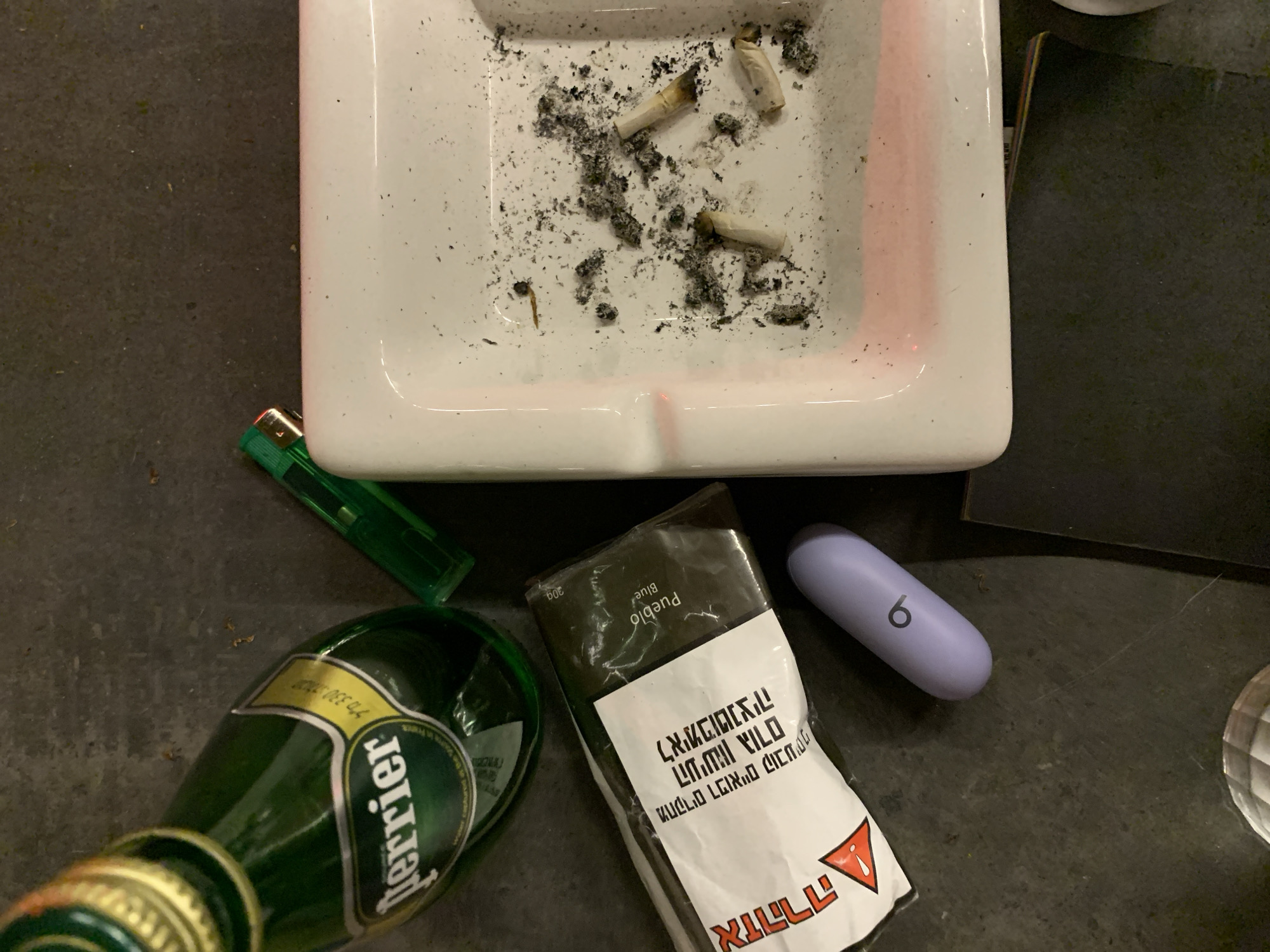 A top-down view presents an arrangement of consumer items on a dark, speckled surface, consistent with a table or countertop, under ambient indoor lighting, situated in Tel-Aviv, Israel.
To the upper left, a green glass bottle of Perrier sparkling water, approximately 330ml, lies on its side, displaying its gold cap and a yellow label with "Perrier" in green text, Hebrew text ("מים מינרליים טבעיים"), and English text indicating its French origin. Adjacent to the bottle is a green plastic disposable lighter.
Below the Perrier bottle and lighter is a package of "Pueblo Blue" rolling tobacco, marked 30g. The white section of its packaging features a prominent red triangular warning symbol and Hebrew text, including "אזהרה: העישון מזיק לבריאות" (Warning: Smoking is harmful to health).
Next to the tobacco package, a light purple, oval-shaped case, bearing a stylized "b" logo, is present, consistent with a wireless earbud case.
Occupying the right side of the frame is a white, square ceramic ashtray. It contains multiple cigarette butts, significant amounts of dark grey ash, and scattered tobacco fragments, indicating recent smoking activity. A faint pink light reflection is visible along the interior edge of the ashtray.
No individuals are visible in the scene. The items suggest recent use by an unseen person.