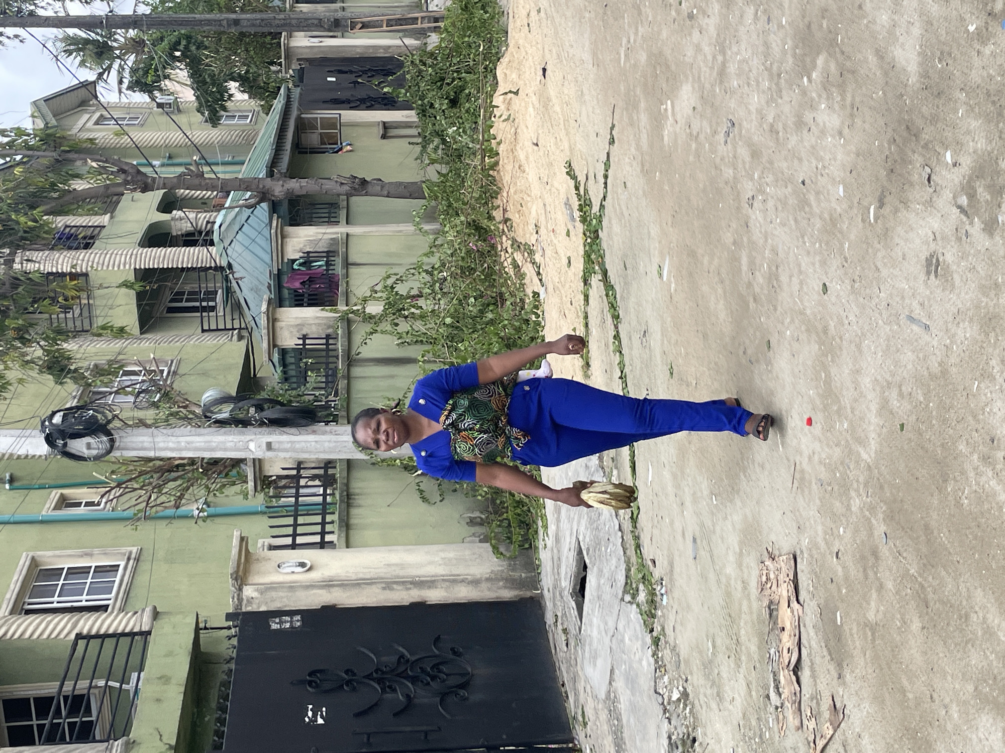 A woman in a royal blue pantsuit stands in a concrete area in Ado, Nigeria. She holds a cluster of bananas. Behind her is a multi-story green building with dark metal gates. Trees and vines grow along the side of the building and onto the concrete. Electrical wires are visible near the top of the building.