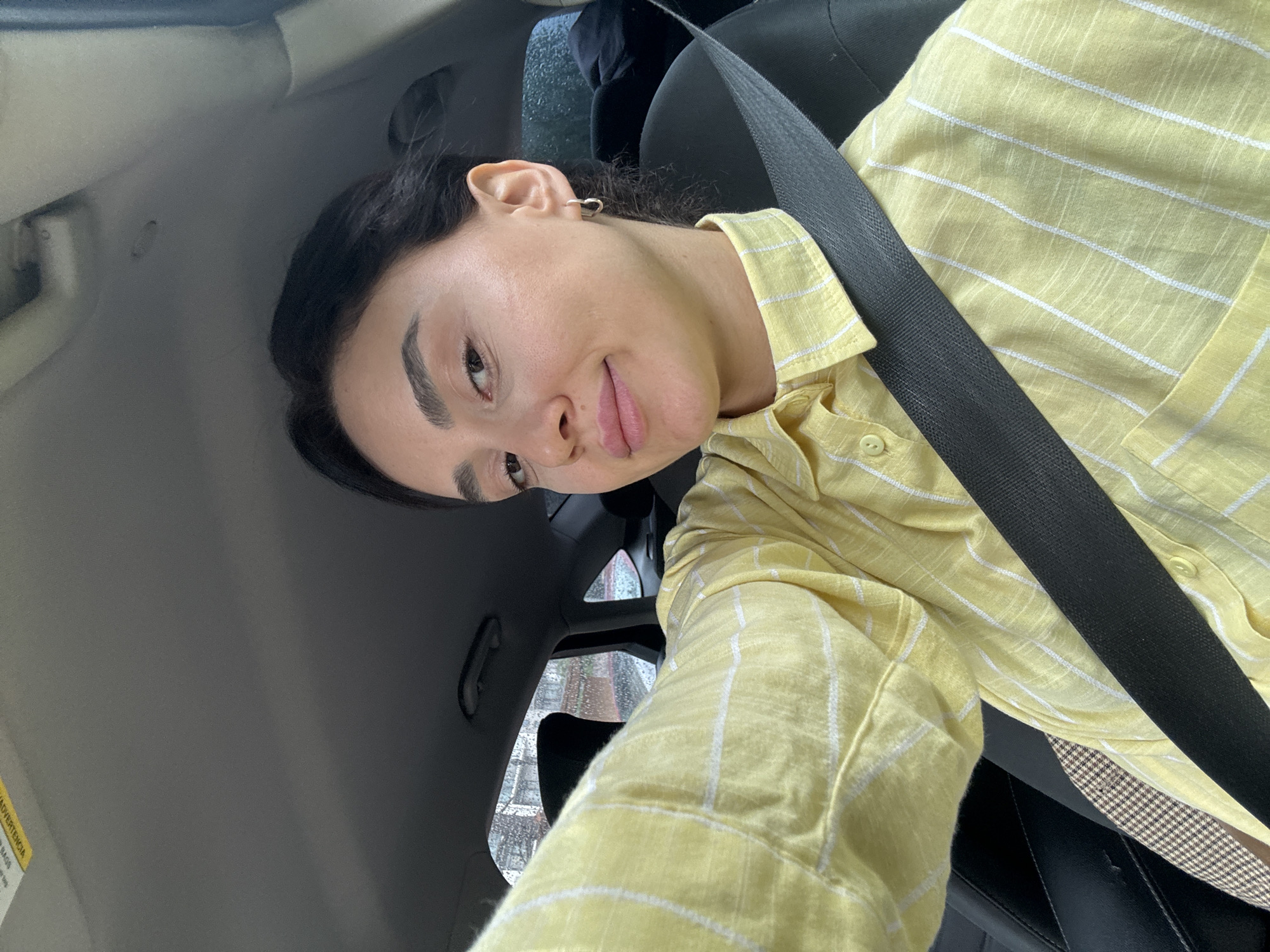 The image depicts a woman in a car, wearing a yellow and white striped shirt and a seatbelt. She has dark hair pulled back, distinct eyebrows, and a heart-shaped earring in her left ear. The background includes the car's interior, with the headrest and window visible. Based on the external view through the window, the location appears to be a street in Tbilisi, Georgia, indicated by unique architectural details and precipitation on the window. The woman is likely sitting in the passenger seat, as she's wearing a seatbelt, and the angle of the shot suggests she's holding the camera. The environment is urban, with possible buildings visible through the rainy window.
