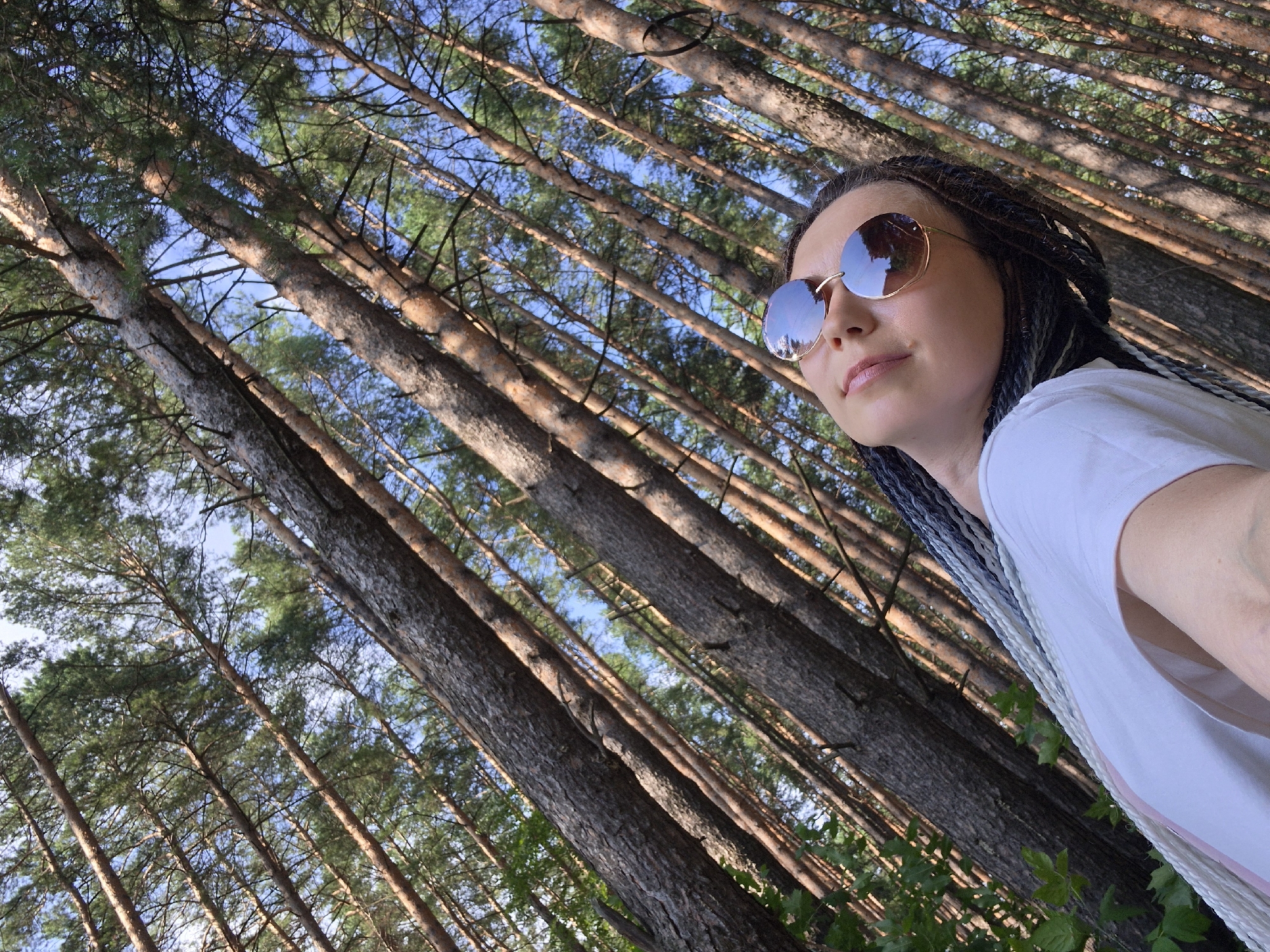 The image shows a low-angle shot of a person with braided hair wearing sunglasses and a white t-shirt in a forest. The background consists of numerous tall pine trees with a blue sky visible through the branches. The person's face is tilted upwards, and their eyes are closed or looking up. The overall scene depicts a person enjoying nature in a forest setting, possibly on a sunny day. The image does not provide specific details about the city of Chaikovsky, Russia.